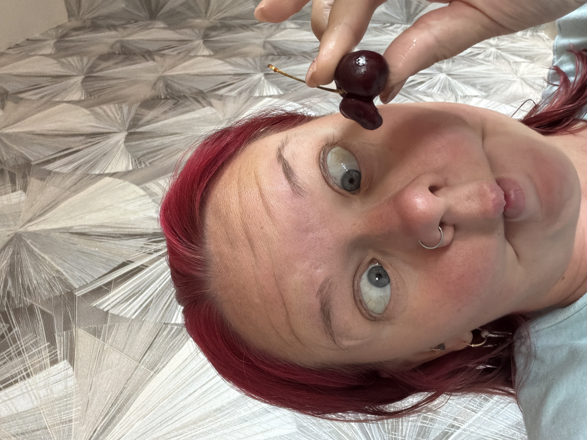 The image shows a close-up of a person with bright red hair and a light blue t-shirt. The person is making a funny face, with crossed eyes and a puckered mouth. They are holding a cherry with two lobes near their face, positioning it in front of their right eye. The background is a patterned wallpaper with abstract, geometric shapes in shades of grey and white. The lighting is bright, suggesting it is daytime. The image appears to be a selfie taken indoors. The location is stated to be in the city of Chaikovsky, Russia.