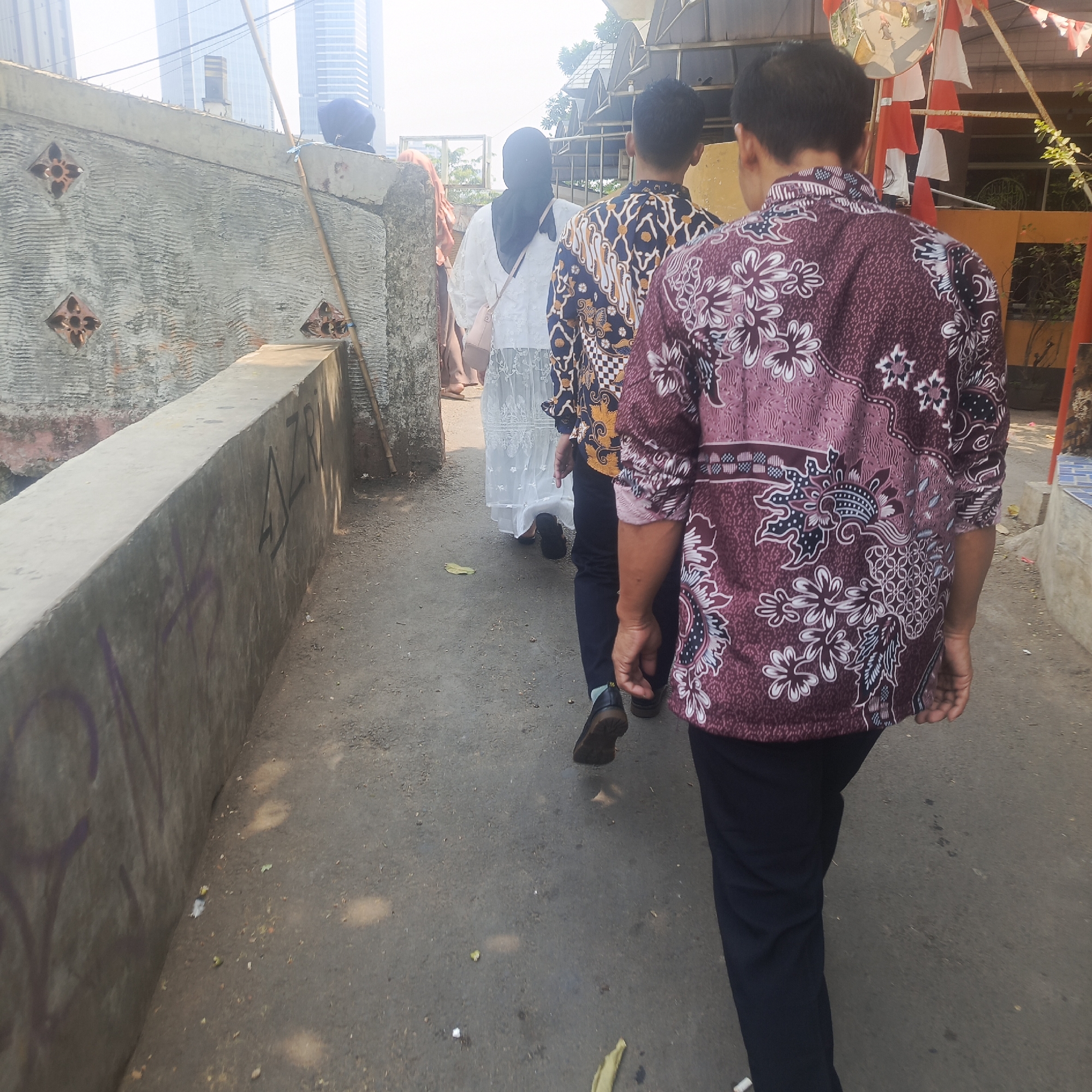In South Jakarta, Indonesia, a group of individuals walks down a narrow paved path next to a concrete wall. The people are dressed in traditional Indonesian batik shirts and modest clothing, with one person wearing a black headscarf and a white dress. The wall to the left has graffiti and decorative tiles. In the background, tall modern buildings contrast with the more traditional surroundings. Red and white Indonesian flags are visible in the upper right corner, suggesting a possible local celebration or event. The lighting indicates it is daytime, with sunlight casting shadows on the ground.