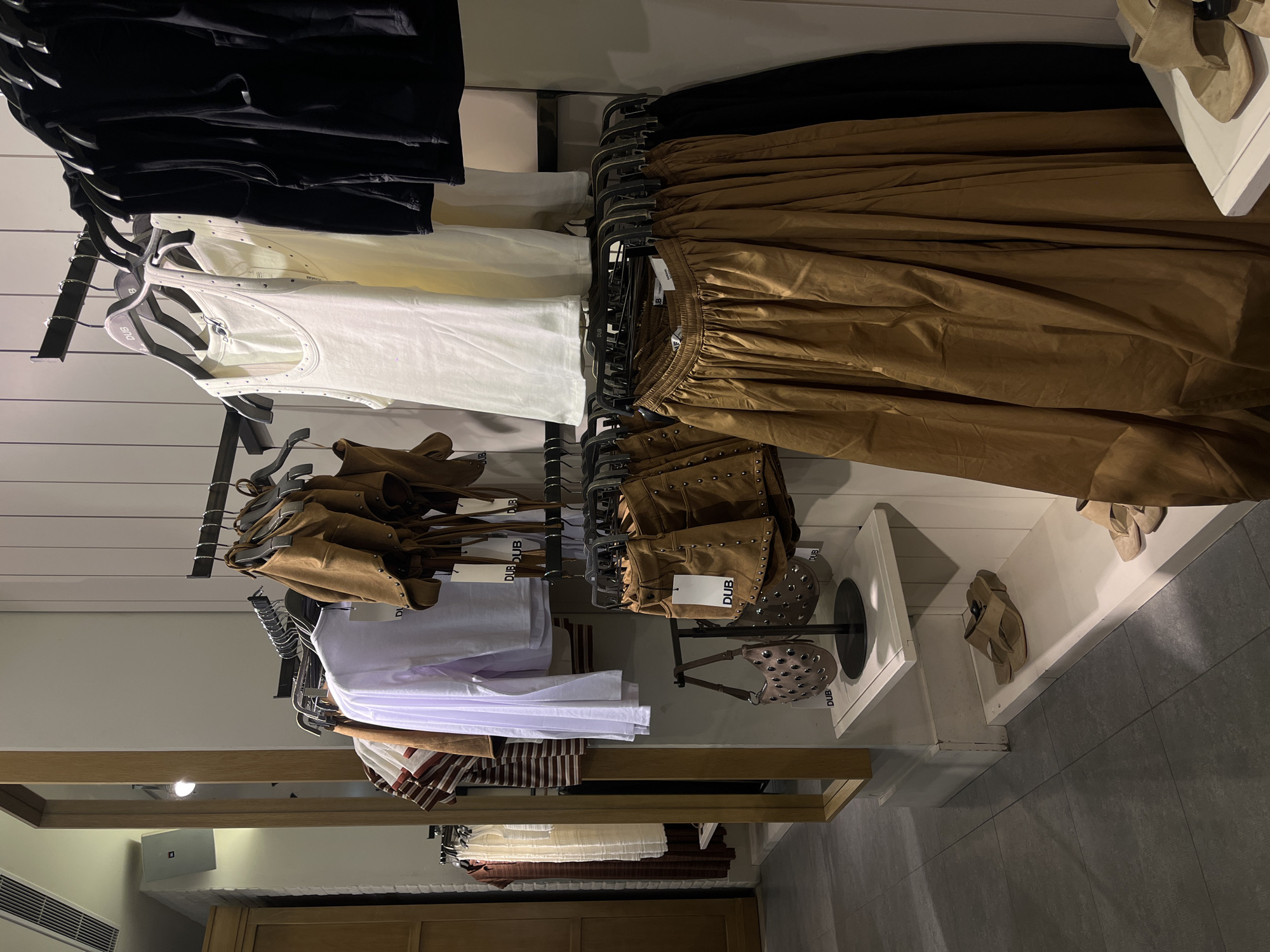 The image captures a retail display within a clothing store in Krasnodar, Russia. The scene showcases various women's garments and accessories arranged on racks and shelves against white slatted walls and wooden fixtures.
On the upper left, black tops or skirts are visible. Below them, white and off-white tank tops, featuring small studs along their edges, hang on dark hangers, some with "DUB" branding. To the right, a rack displays numerous pairs of light brown or tan wide-legged trousers with elasticated waists.
Further down, on the left, folded white shirts and striped tops in white and brown are presented. Adjacent to these, more studded brown or tan garments, possibly shorts or skirts, are hung individually, also bearing "DUB" tags. A small, studded brown handbag is displayed on a stand nearby.
On the right side of the image, light beige or tan shoes/sandals are placed on white display shelves. The floor is covered with grey tiles. The store interior features artificial lighting, an air conditioning vent on the far left, and a JBL speaker mounted on a wall in the background. The overall presentation is organized, with a dominant neutral color palette of black, white, and various shades of brown and tan.