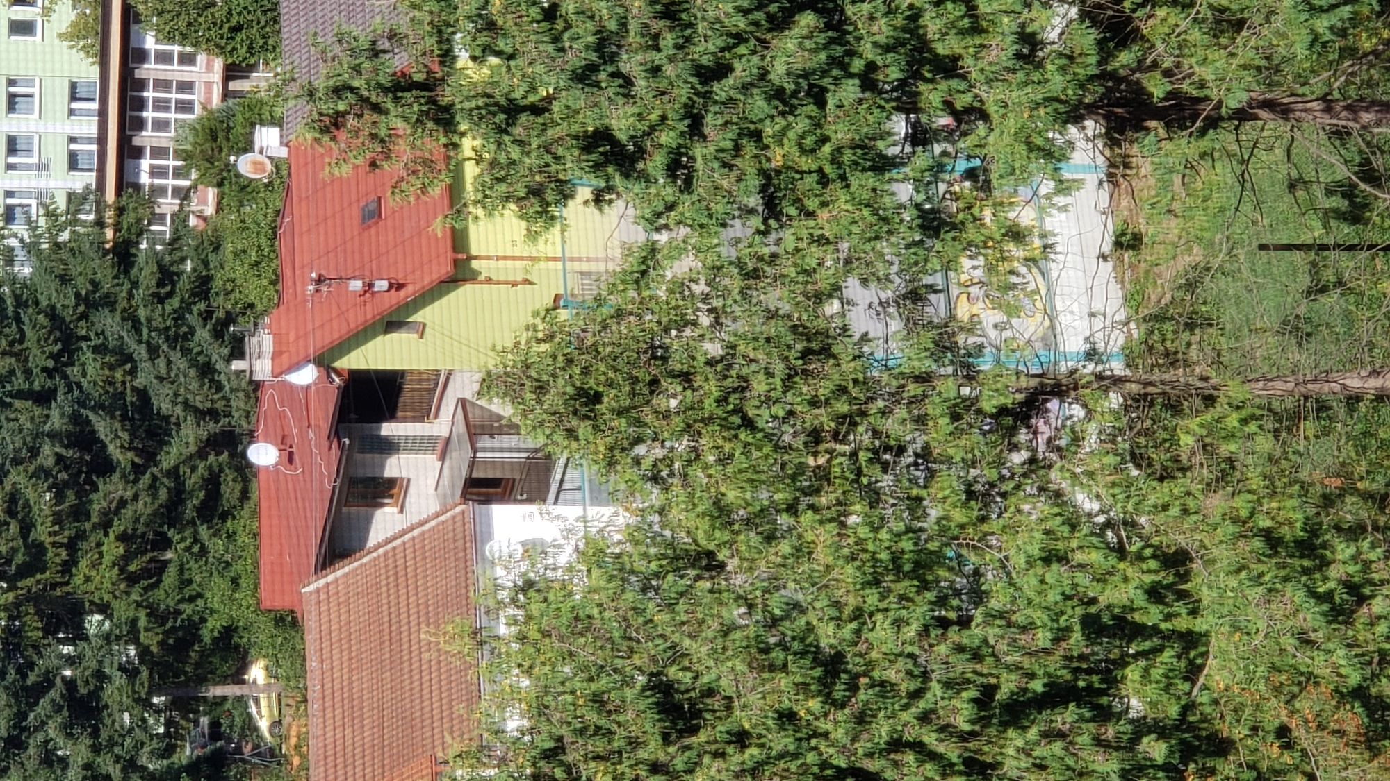 The image shows a residential area with several houses and apartment buildings partially obscured by lush green trees. The buildings have red and light green roofs, some with satellite dishes. There is a yellow car visible on a street in the background, and various architectural features like windows, balconies, and chimneys. The setting appears to be a sunny day with shadows cast by the trees, suggesting an outdoor environment. The buildings and foliage indicate a suburban or semi-urban landscape. The image lacks any people or discernible interactions.