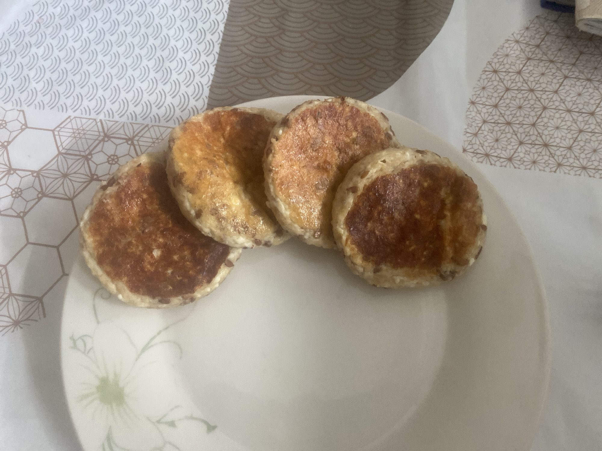 The image shows four round, browned patties arranged on a white plate with a floral design. The plate rests on a patterned tablecloth with white and beige tones. The patties appear to be some kind of food, possibly small pancakes or fried cakes, with a browned or slightly burnt surface. The scene is indoors with natural lighting, but there is no information about the location in Budva, Montenegro. No people, actions, or interactions are visible.