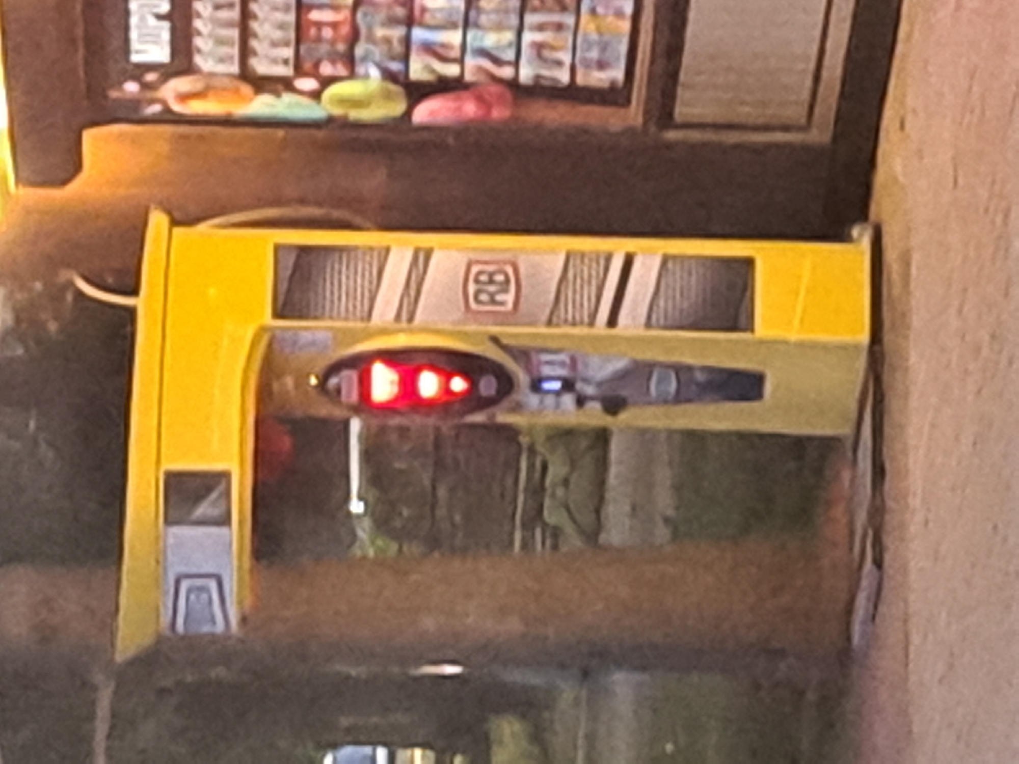 The image depicts a bright yellow vending machine with red lights and the letters "RB" visible on its side, located in Irkutsk, Russia. Adjacent to the vending machine is a display of snacks and drinks. The scene appears to be outdoors, with trees and a sidewalk visible in the background. The lighting suggests it might be dusk or nighttime.