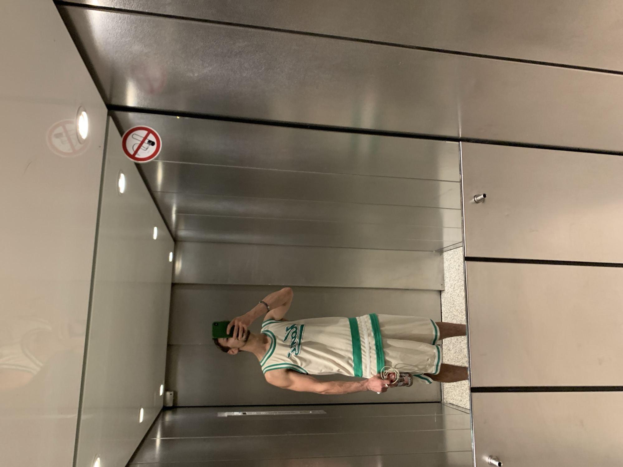 A person is taking a mirror selfie in what appears to be a modern elevator. The person is wearing a white and green basketball jersey and shorts. They are holding a can in their left hand and a cell phone in their right hand. The elevator walls are made of brushed metal. A "no smoking" sign is visible on the ceiling. The floor of the elevator is speckled and light-colored. The image is taken in Worms, Germany.