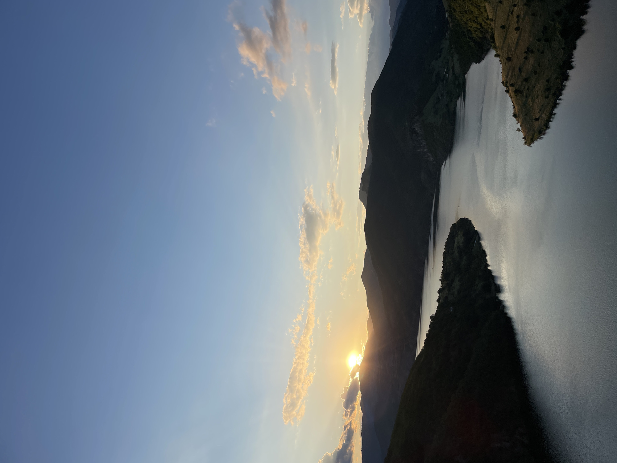 This is a landscape photograph of a scenic view near Borovitsa, Bulgaria, captured during sunset. The image features a wide, calm body of water, likely a reservoir, winding through a valley surrounded by dark, silhouetted mountains. The setting sun is partially obscured by the mountain range on the left side of the frame, casting warm golden light and long shadows across the scene. Wispy clouds tinged with orange and pink are scattered across the upper portion of the sky, transitioning to a clear blue towards the zenith. The slopes of the mountains are covered in vegetation, with some areas showing grassy hillsides in the foreground and darker, forested slopes further back. The overall atmosphere is serene and picturesque, highlighting the natural beauty of the Bulgarian landscape at dusk.