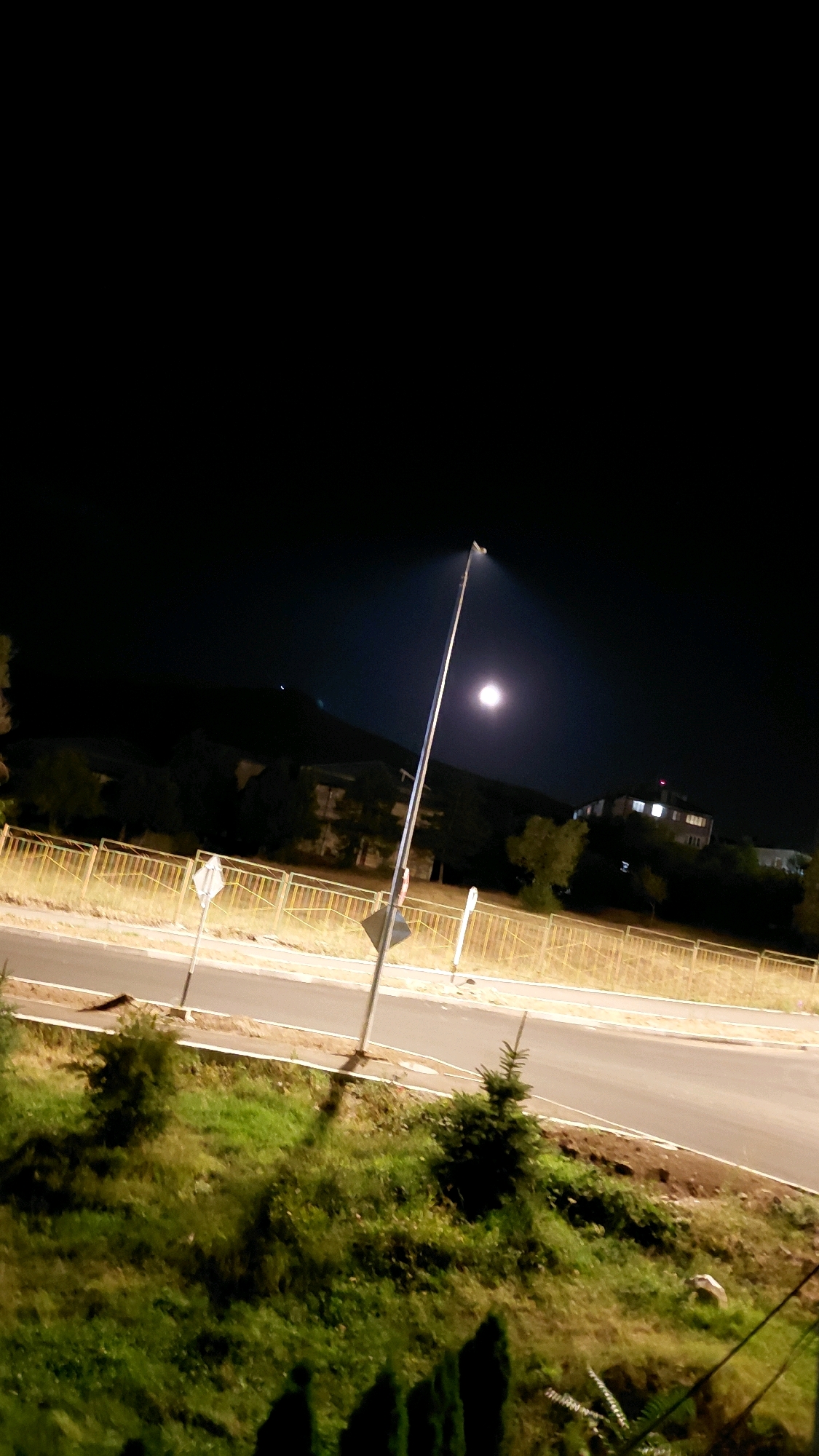 The image captures a night scene in the city of Mayakovski, Armenia, focusing on a street illuminated by a lone street lamp. A full moon is visible in the dark sky, casting a bright glow. The street itself is paved and appears to be an urban road, with a yellow metal fence running alongside it. Beyond the fence, there is a grassy embankment with scattered trees and bushes. In the background, several buildings are visible, some with lights on, suggesting they are inhabited or operational. The overall atmosphere is one of quiet nighttime urban tranquility.