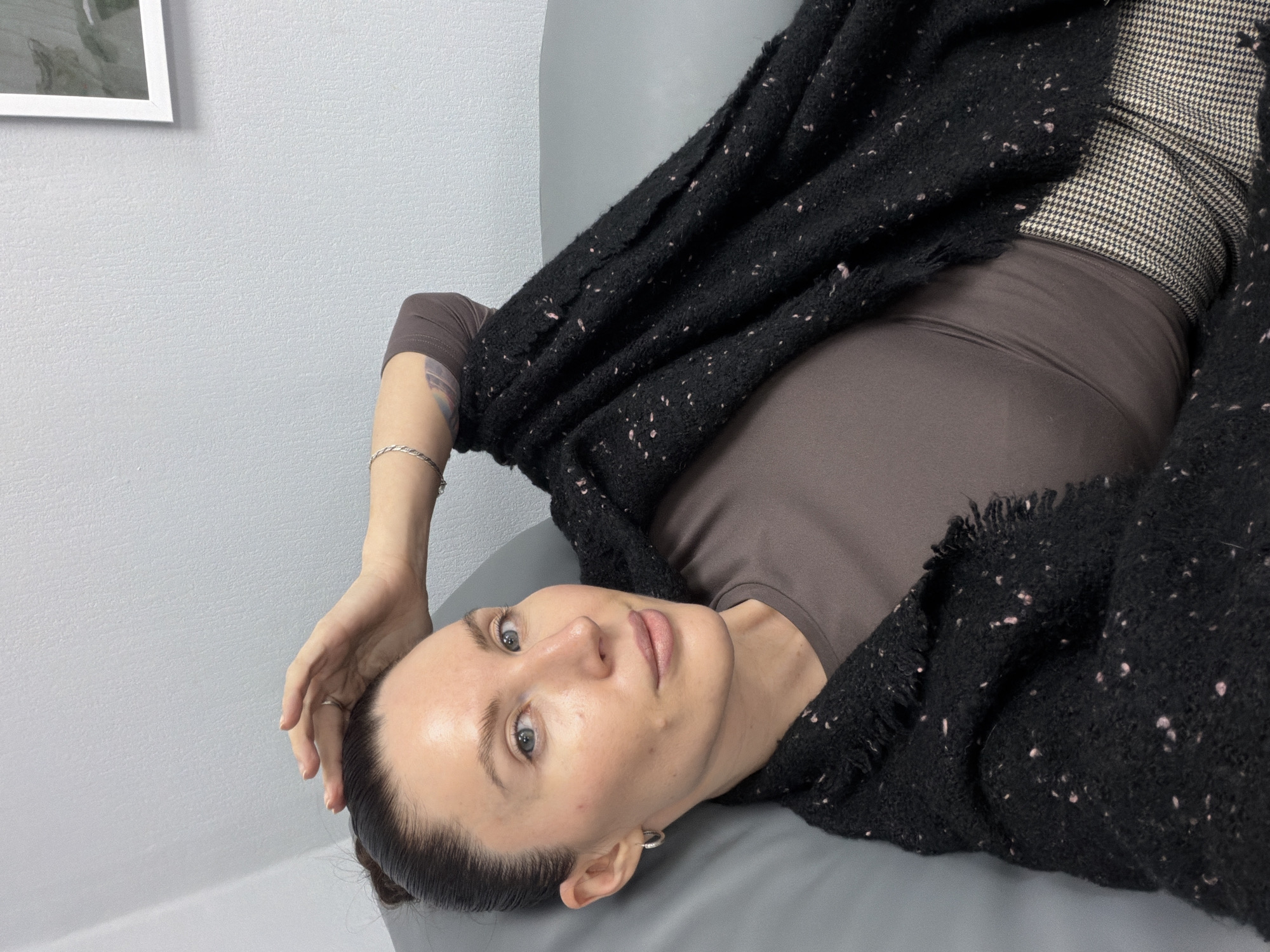 A woman lies on a gray surface in an indoor setting. She wears a brown long-sleeved shirt, houndstooth patterned pants, and a black shawl. Her right arm is raised above her head, and she looks towards the camera. The background is a light gray wall with a framed picture hanging on it. No information about the location in Perm, Russia, can be determined from the image.