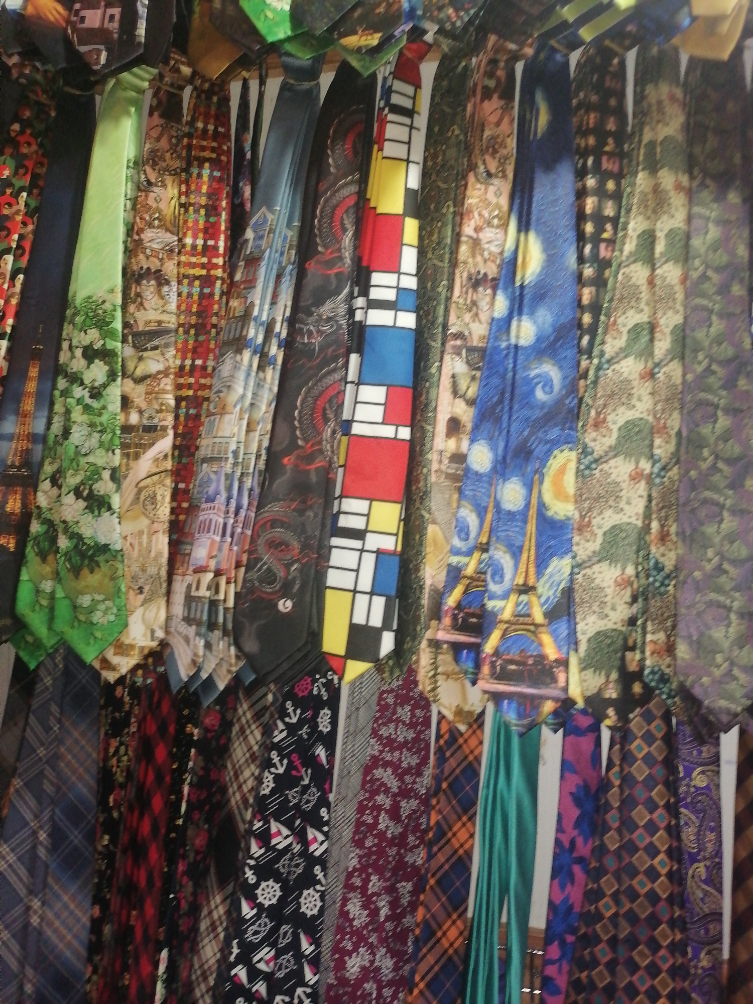 The image depicts a vibrant and colorful display of neckties, arranged in a vertical manner. The ties exhibit a wide variety of patterns and designs, including floral motifs, geometric shapes, cityscapes, and abstract art. Some of the ties appear to be made of silk, given their sheen and drape.
The background suggests a retail environment, possibly a market stall or a shop. The lighting is somewhat dim, but adequate to illuminate the individual ties. The overall impression is one of abundance and variety, catering to diverse tastes.
This scene is likely located in a bustling commercial area of Lagos, Nigeria, a city known for its dynamic markets and rich textile culture. Such a display of neckties would be fitting for an establishment selling fashion accessories in a vibrant Nigerian city.