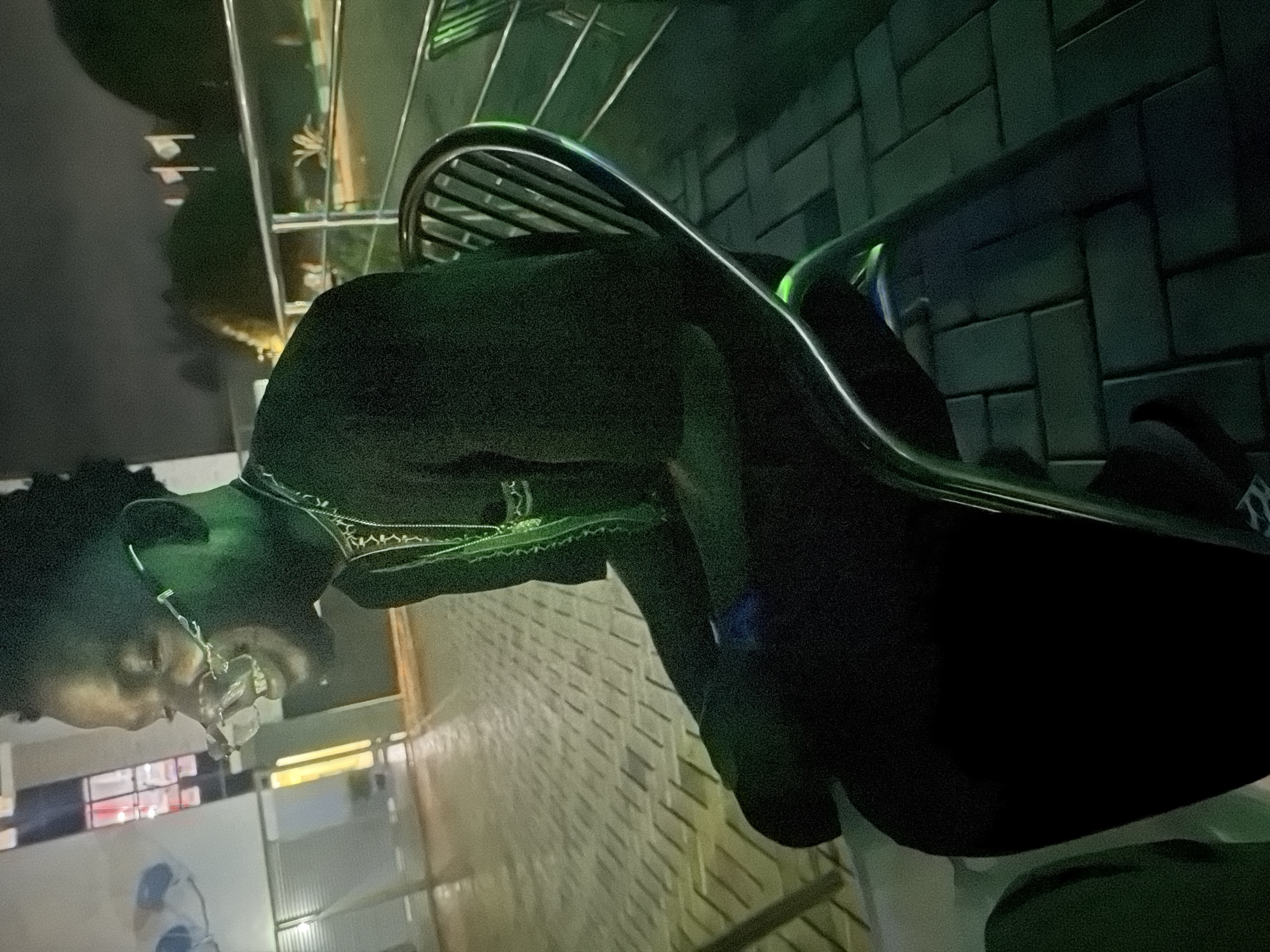 In Bukuru, Nigeria, a young Black male sits on a metallic chair outside at night. He is wearing a black garment with intricate white and green embroidery around the neckline and a silver chain with a pendant. He has on distinctive eyeglasses with angular frames. The man is facing left and smiling, his mouth open, revealing his teeth. The background is dimly lit, with paving stones visible on the ground, suggesting an outdoor seating area. A metal railing and more chairs are visible in the background, hinting at a public space or a restaurant. The lighting casts a green hue over the scene, likely from artificial sources.
