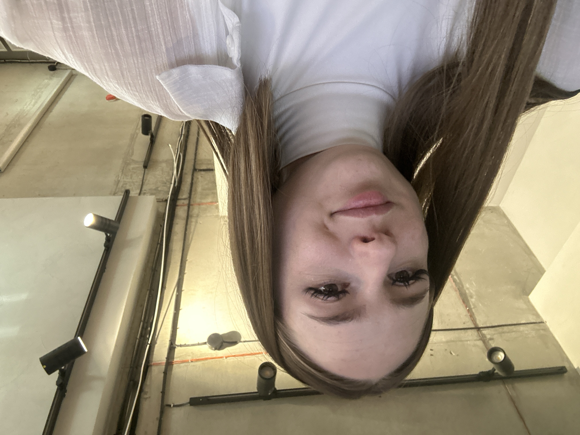 A young Caucasian woman with long, brown hair and prominent eyelashes takes a selfie. She is wearing a white turtleneck shirt and a light-colored, sheer collared shirt over it. The background shows a concrete ceiling with exposed pipes and track lighting, suggesting an indoor, possibly industrial or modern architectural setting. The lighting is bright, casting some shadows.