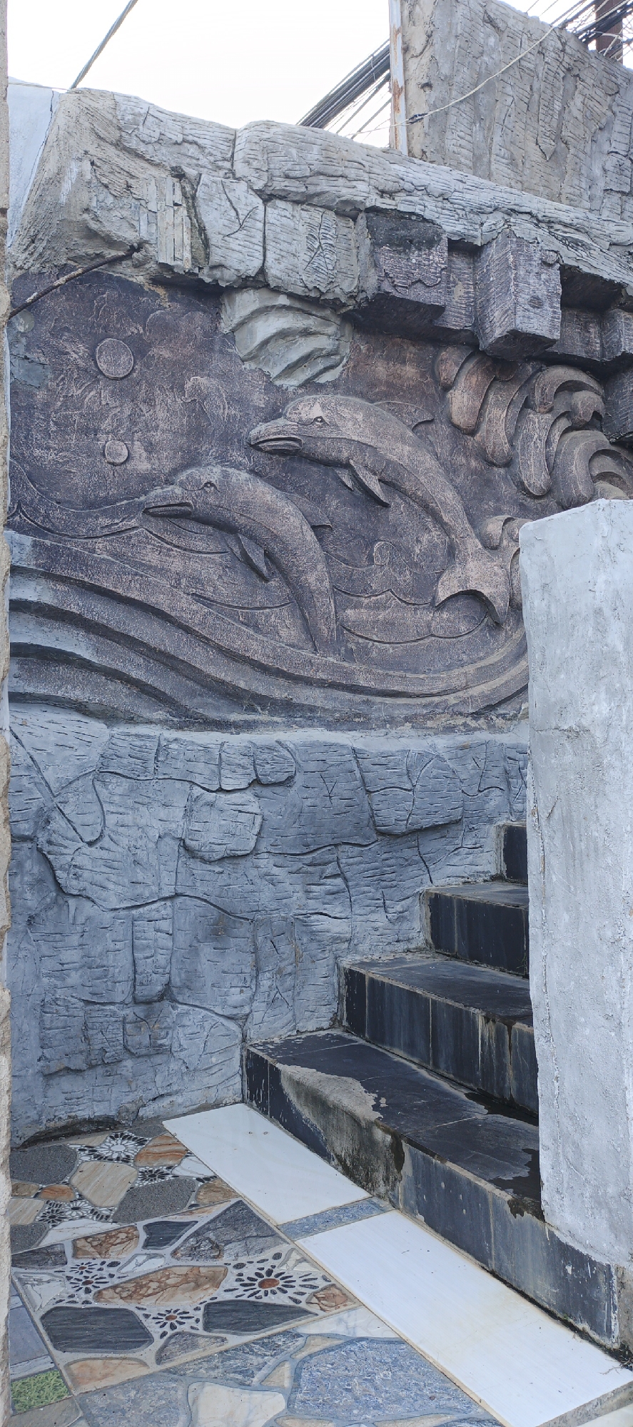 This is a full-shot, eye-level, outdoor photograph of a decorative wall with a dolphin relief and a set of stairs. The wall, made of concrete, features a textured relief depicting two dolphins swimming in wavy water. Above the dolphins, there are two circular shapes that may represent the moon or sun. The wall itself is designed to resemble layered rock formations, with a rough, grey upper section and a darker, textured section for the dolphin relief. Below the relief, the wall continues with a grey, rocky texture. To the right of the wall, a set of dark grey stairs with a rough finish ascends. The steps appear wet and slightly worn. The ground in front of the stairs is tiled with a mosaic of irregular shapes in various earthy tones, with some tiles featuring floral patterns. A light grey concrete pillar is visible to the right of the stairs. The background shows a neutral sky and some wires overhead, suggesting an urban or semi-urban environment. The scene is located in Phu Thuy Ward, Vietnam. There are no people visible in the image.