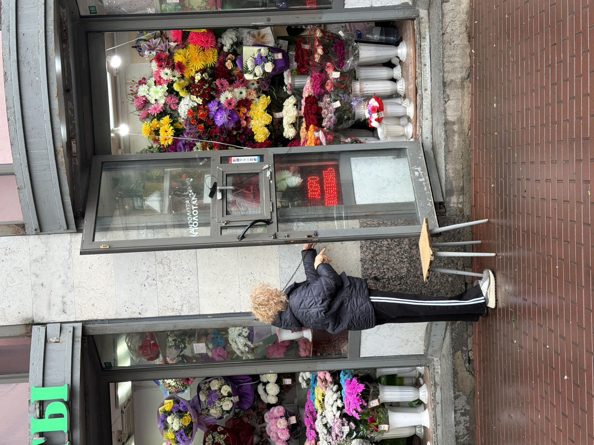 A woman stands outside a flower shop. The shop has a large window display filled with a variety of colorful flowers and bouquets. The woman is wearing a black quilted jacket and black pants with a white stripe. She is reaching up to adjust or interact with the shop's entrance. A small, rudimentary stool sits on the ground next to her. The ground is a mix of red brick and stone. A green sign is visible on the left side of the image. Based on the architectural style and signage, the location is Saint Petersburg, Russia.