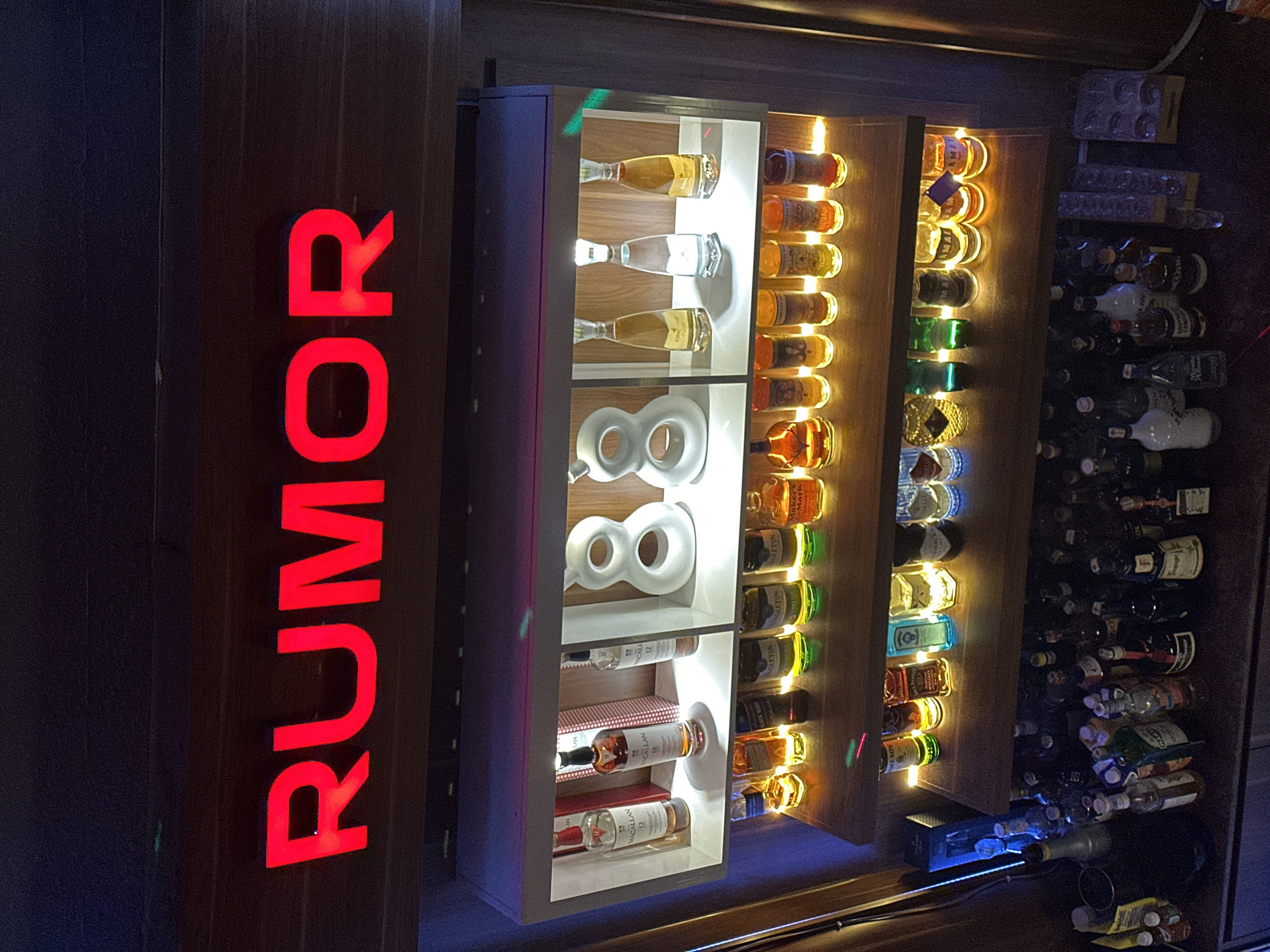 In Khan Chamkar Mon, Cambodia, a dimly lit establishment features a prominent "RUMOR" sign in large, red, illuminated letters. Below the sign, a multi-tiered display shelf showcases a variety of liquor bottles and decorative items. The top shelf has three bottles of Macallan Scotch whisky and two white, sculpted "8" shapes. The second shelf displays numerous small liquor bottles, some with yellow or green liquid, illuminated by soft lights. The third shelf contains more small bottles, including some with colored liquid. The lowest shelf, not fully visible, appears to hold a collection of larger liquor bottles and glassware. The environment suggests a bar or liquor store setting, with wooden paneling and ambient lighting contributing to a sophisticated or atmospheric ambiance.