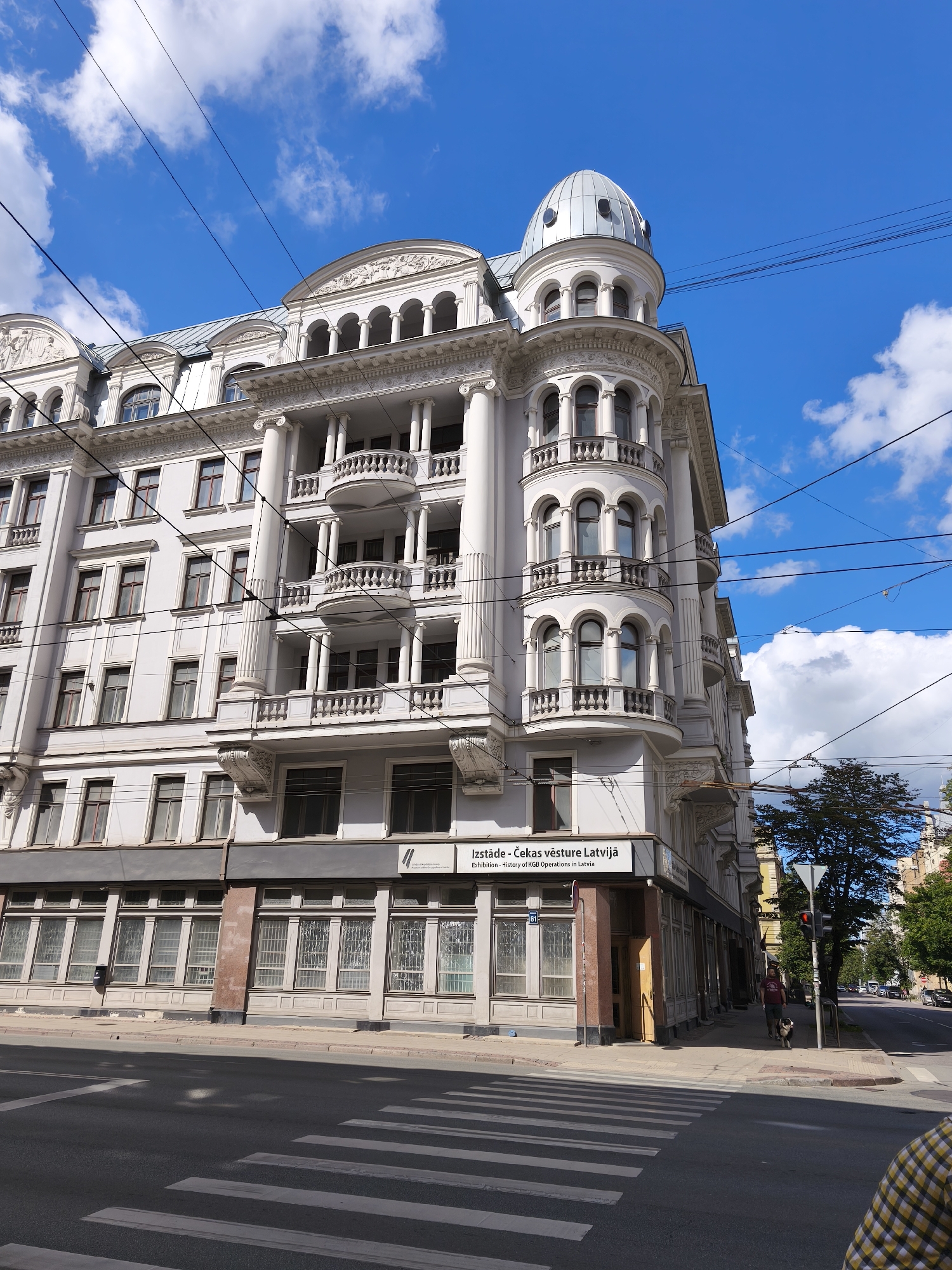 The image shows the exterior of a building located at Brīvības iela 61 in Riga, Latvia. This building houses the "Izstāde - Čekas vēsture Latvijā" (Exhibition - History of KGB Operations in Latvia). The architecture is ornate, featuring classical elements like columns, arched windows, and balconies, with a distinctive dome atop a corner turret. The building is a light color, possibly white or pale gray, and is multi-storied.
In the foreground, a crosswalk is visible on an asphalt road, indicating a pedestrian crossing area. The sky is blue with scattered white clouds. Overhead, a complex network of electrical wires crisscrosses the scene, typical of urban environments.
On the sidewalk in front of the building, a person is walking a dog. The person is wearing a dark t-shirt and shorts. The dog appears to be medium-sized. Further down the street, more buildings and trees are visible, as well as traffic signals and what appear to be other pedestrians. The overall atmosphere suggests a sunny day in a city.