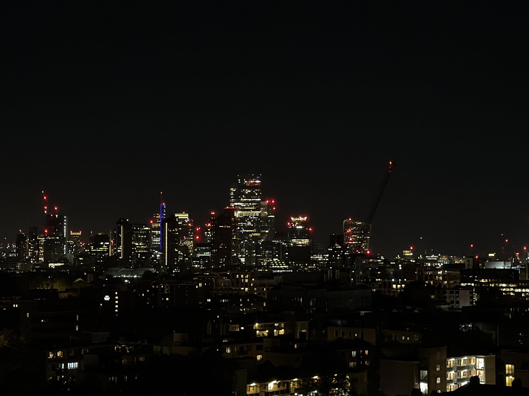 This is a nighttime cityscape of London, United Kingdom. The scene shows a skyline of illuminated buildings of varying heights and shapes, indicating an urban environment. Lights from the buildings, streetlights, and possibly vehicles create a network of brightness against the dark sky. The presence of cranes, indicated by red lights at the top, suggests ongoing construction or development projects in the area. A lower layer of smaller buildings or houses can be seen closer to the camera, also with some lights on. The overall impression is a view of a busy and developed urban area at night.
