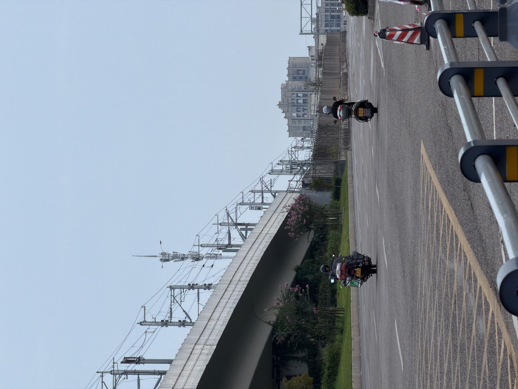 In Jiangjin, China, on an overcast day, two individuals are seen riding motorcycles on a multi-lane paved road. The foreground features a metal railing with yellow accents, a traffic cone with red and white stripes, and yellow and grey painted road markings. In the midground, the two motorcyclists are traveling in the same direction, away from the camera. The rider on the left has a green and red motorcycle with luggage strapped to the back and sides, and appears to be wearing a dark helmet and jacket. The rider on the right is on a dark-colored motorcycle with a passenger seat, and the rider is wearing a grey helmet and a dark top. The background includes a large, elevated concrete structure, possibly a highway or overpass, with numerous overhead electrical wires and poles. Beyond the overpass, a communication tower is visible, and further in the distance, several multi-story buildings form a city skyline. Green vegetation lines the side of the road under the elevated structure.