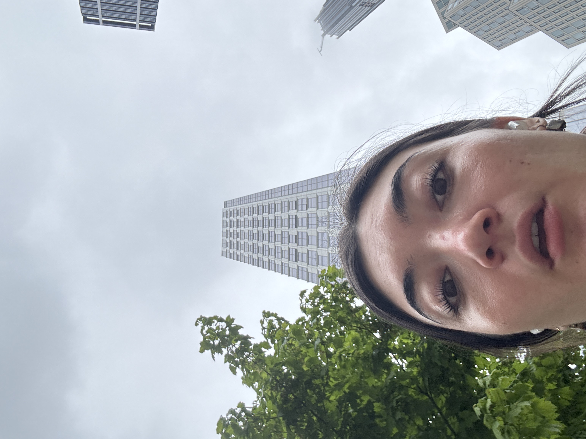 The image presents a low-angle view, predominantly featuring a woman's face in the foreground, set against an urban backdrop with natural elements. The woman's right eye, with prominent eyelashes and dark eyebrows, nose, and partially open mouth are visible on the right side of the frame. Her dark hair is pulled back, with some strands visible near her forehead and ear.
In the background, a cloudy, grey sky dominates the upper and left portions of the image. Several tall, modern skyscrapers are visible, characterized by their numerous windows and vertical lines, suggesting a dense urban environment. One particularly slender building stands centrally in the background. Below the buildings and partially obscuring them on the lower left, lush green tree foliage is visible, indicating the presence of trees or a park area within the cityscape.
The perspective of the shot suggests it was taken from a low angle, possibly a selfie or by someone positioned beneath the subject, looking upwards. The time of day appears to be daytime, under an overcast sky. There is no specific activity depicted other than the woman seemingly posing for the photograph. Her expression is neutral as she looks towards the camera. There is no visible text in the image. The combination of skyscrapers and trees is consistent with a major metropolitan area like New York, United States of America.