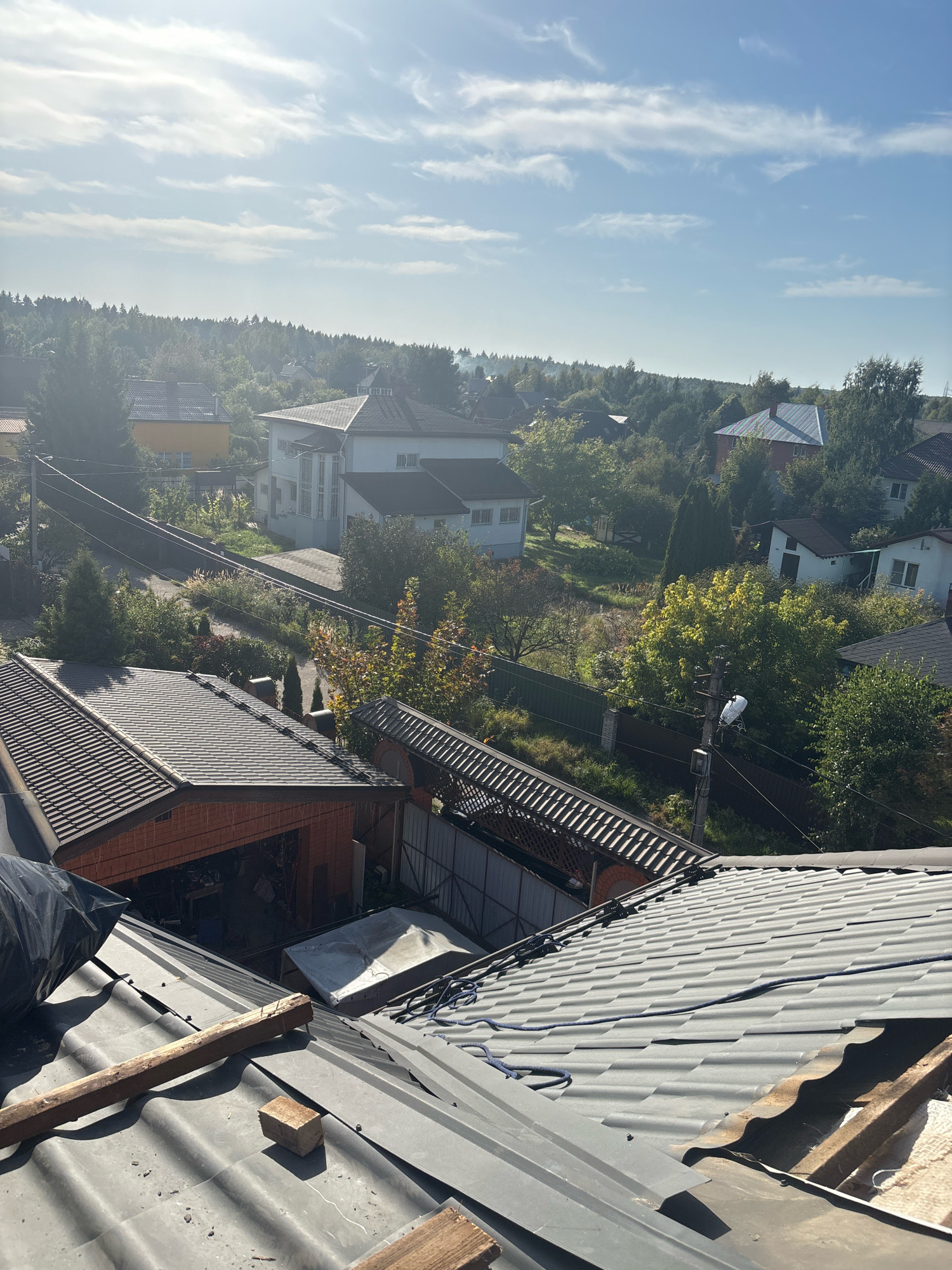 The image shows a panoramic view of a suburban area with houses, trees, and a cloudy sky. The foreground features several rooftops, some covered with dark gray, corrugated metal tiles, and others with a mix of asphalt shingles and exposed wood. Construction materials, including a black plastic tarp and wooden planks, are visible on one of the rooftops, suggesting ongoing renovation or repair work. In the mid-ground, various houses with different roof styles and colors, such as white and yellow, are scattered among lush green trees. Some of the houses appear to be two-story structures with multiple windows. A network of power lines runs across the scene, supported by utility poles. The background is dominated by a dense forest canopy that stretches to the horizon, under a light blue sky with scattered white clouds. The overall impression is that of a peaceful, residential neighborhood nestled within a natural landscape, likely in the city of Zhavoronki, Russia, given the provided context. No people are visible in the image.
