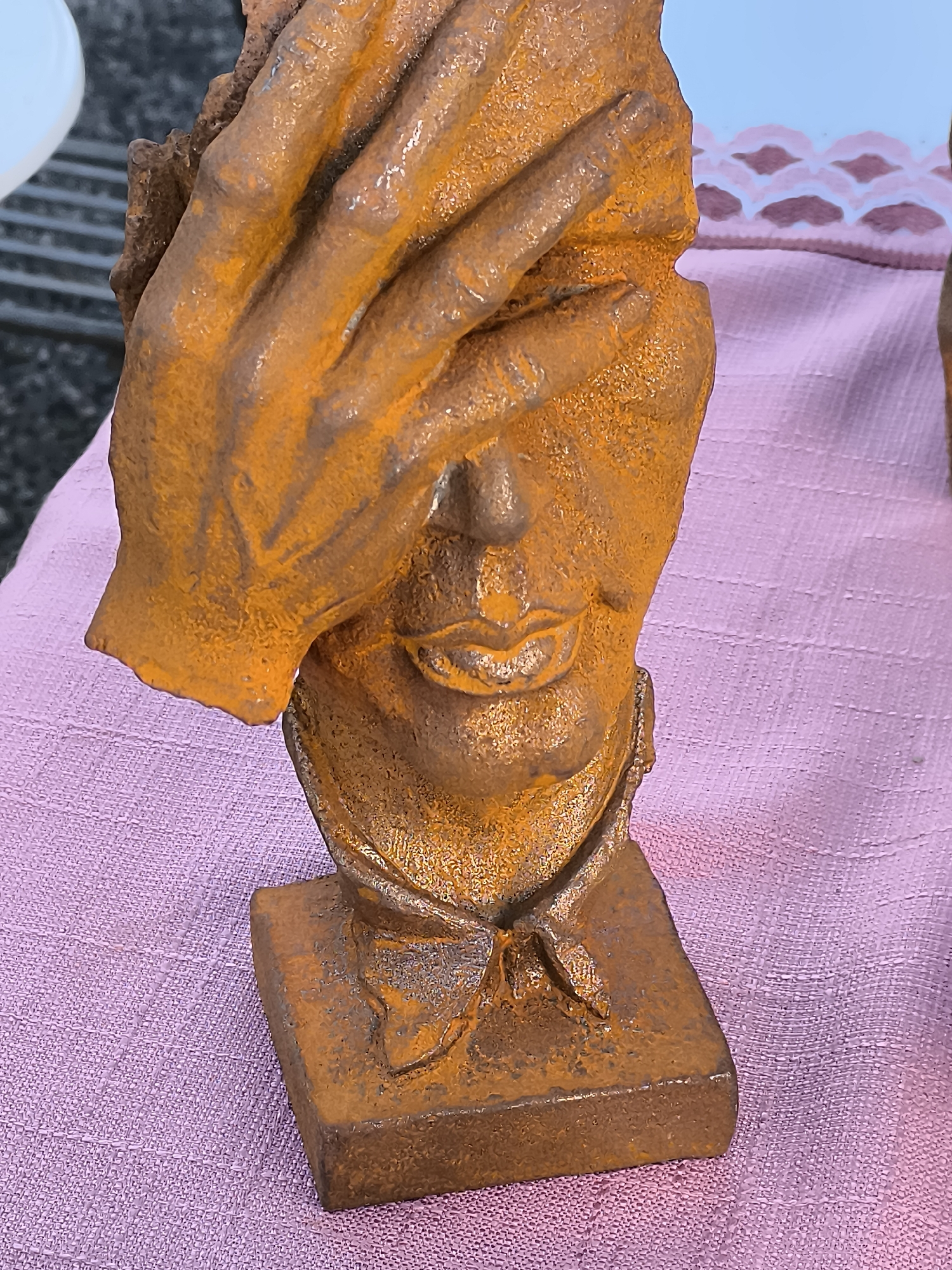 A close-up, eye-level, medium shot features a rusty, orange-brown metal sculpture of a face covered by a hand. The sculpture rests on a square base and is placed on a pink, textured fabric. The fabric appears to be a tablecloth. In the background, to the left, a white, rounded object is partially visible, and beyond that, a dark, slatted surface can be seen. To the right of the sculpture, further back, a decorative pattern of pink and white diamonds on a fabric is visible. The sculpture's surface is rough and pitted, with a patina of rust. The hand is positioned over the eyes of the face, and the mouth is slightly open, revealing a hint of teeth. A small, bow-like tie is sculpted around the neck area of the face. The lighting is somewhat diffused, casting subtle shadows on the sculpture and fabric. The overall impression is of an artistic piece displayed outdoors, possibly at a market or fair in Klaipėda, Lithuania, given the context.