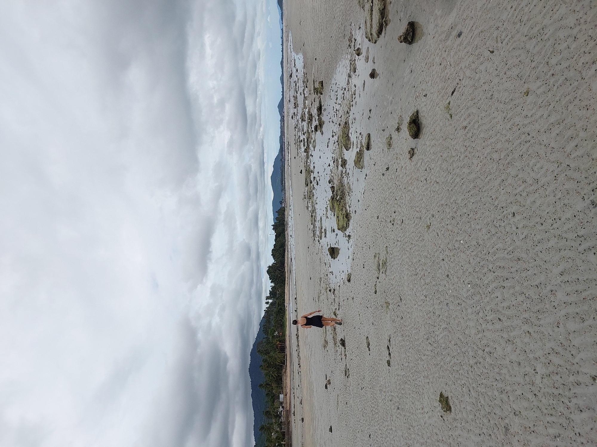 A woman in a black dress walks on a sandy beach under a cloudy sky. The beach is covered with small rocks and shallow pools of water. In the background, there are palm trees and mountains. The scene appears to be in the Ko Samui District of Thailand.