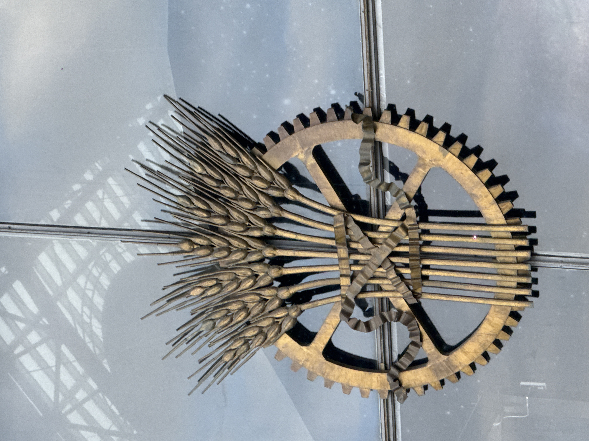 This image shows a metal sculpture featuring a gear, sheaves of wheat, and what appears to be a ribbon. The sculpture is attached to a structure with glass panels. The sculpture is located in Moscow, Russia, and is part of a ceiling decoration. It's a symbol referencing industry and agriculture, possibly representing a period of the Soviet Union. There are no people depicted.