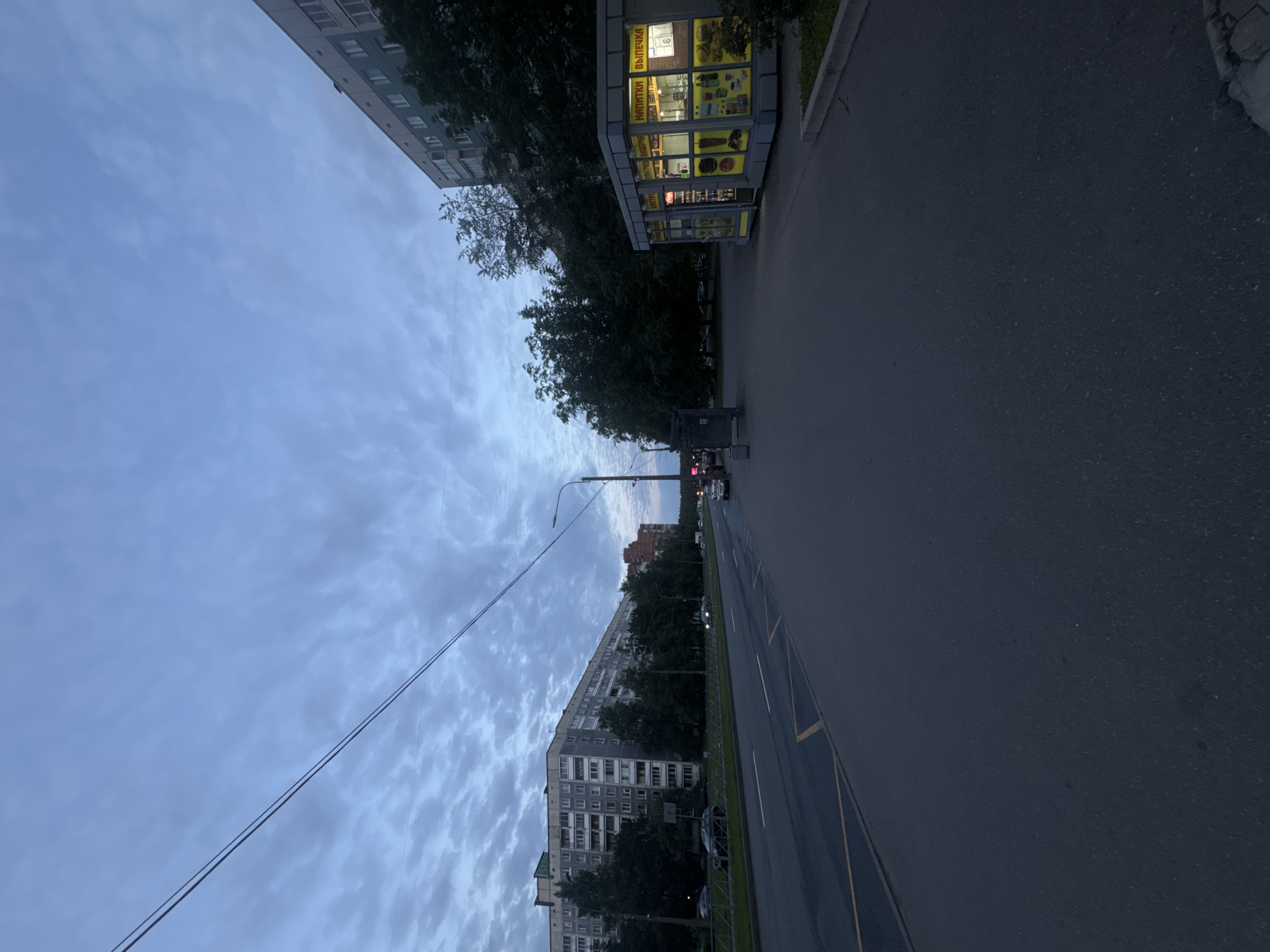 The image depicts a street scene in Sinyavino, Russia, under a cloudy twilight sky. On the left, a long, multi-story apartment building with many windows lines the street. Across the road, a smaller, modern kiosk is illuminated with bright yellow lights, displaying advertisements for "НАПИТКИ" (drinks) and "ВЫПЕЧКА" (baked goods). Trees are visible along the street, and power lines stretch across the sky. Several cars are parked or driving on the road, and a pedestrian walkway is visible in the foreground. The overall impression is a quiet, early evening in a residential urban area.