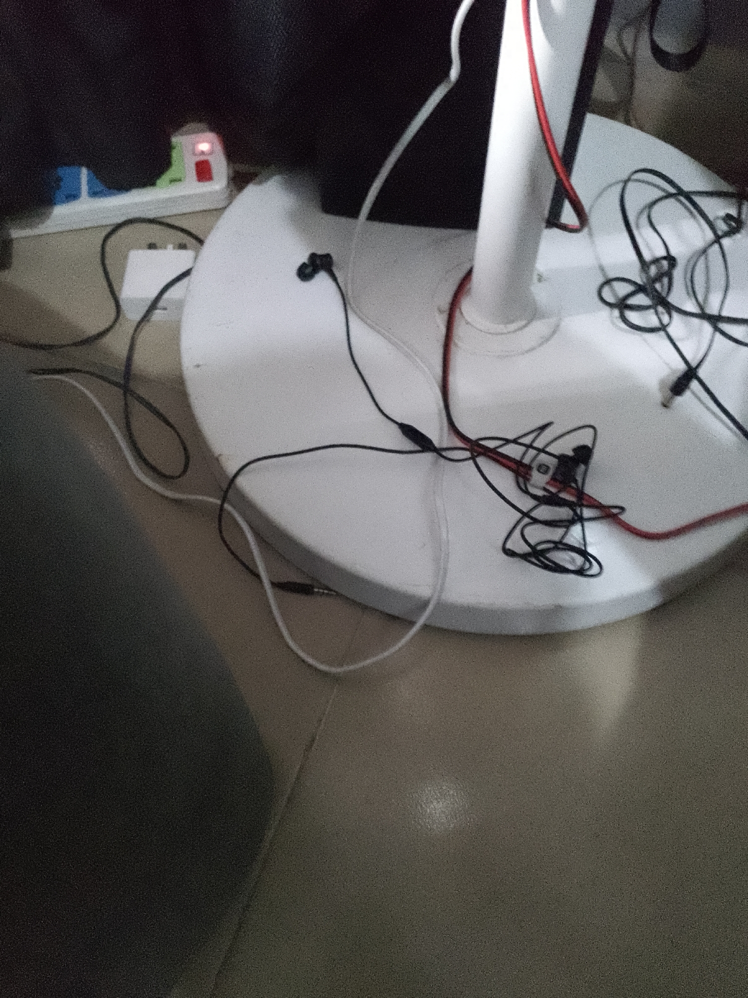 The image shows a white, circular fan base with several black and red wires and cables tangled on its surface. Some of the wires are connected to small black earbud headphones. To the left of the fan base, a white power strip with multiple outlets is visible, with a red indicator light illuminated. One black wire from the fan base appears to connect to a small white adapter box placed on the floor. The fan base and surrounding floor are light-colored. The background is dark and out of focus. The scene appears to be indoors, likely in a home or office environment.