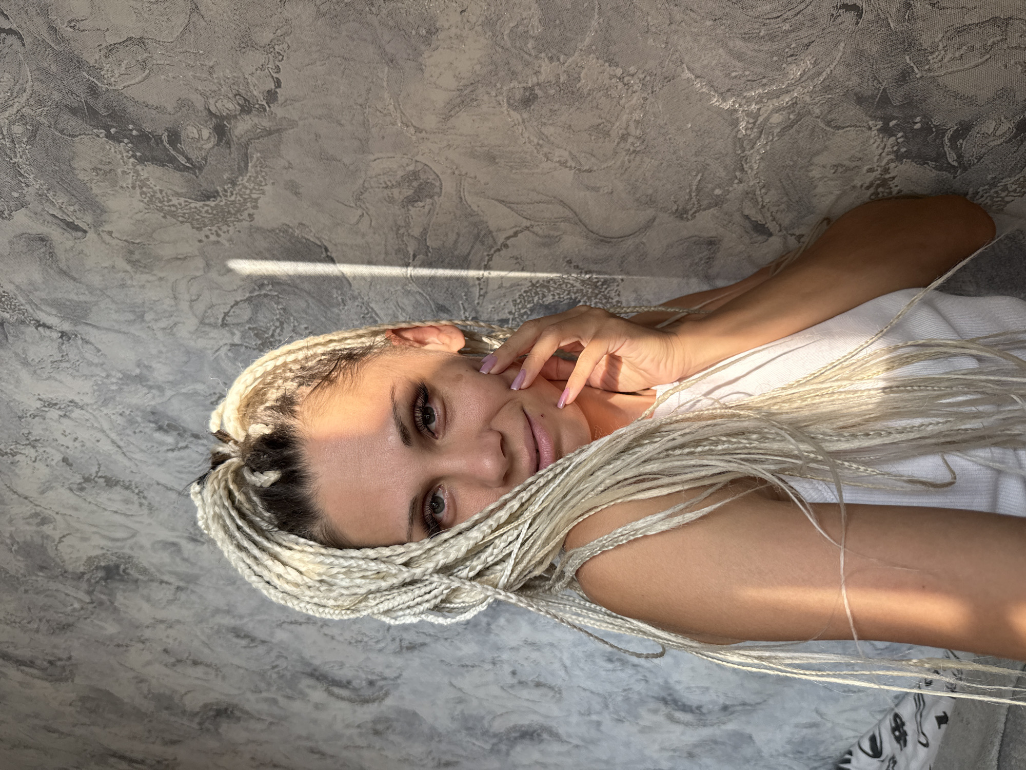 The image is a portrait of a woman with blonde dreadlocks, wearing a white tank top. She is positioned in front of a textured grey wall, with a ray of sunlight casting a shadow diagonally across her face and left shoulder. Her right hand is gently touching her cheek, and her fingernails are painted a light purple. The background appears to be an interior wall with a marbled or distressed pattern. The image was taken indoors, likely in a residential setting, but there is no information provided to determine a specific location within Rusanovo, Russia.