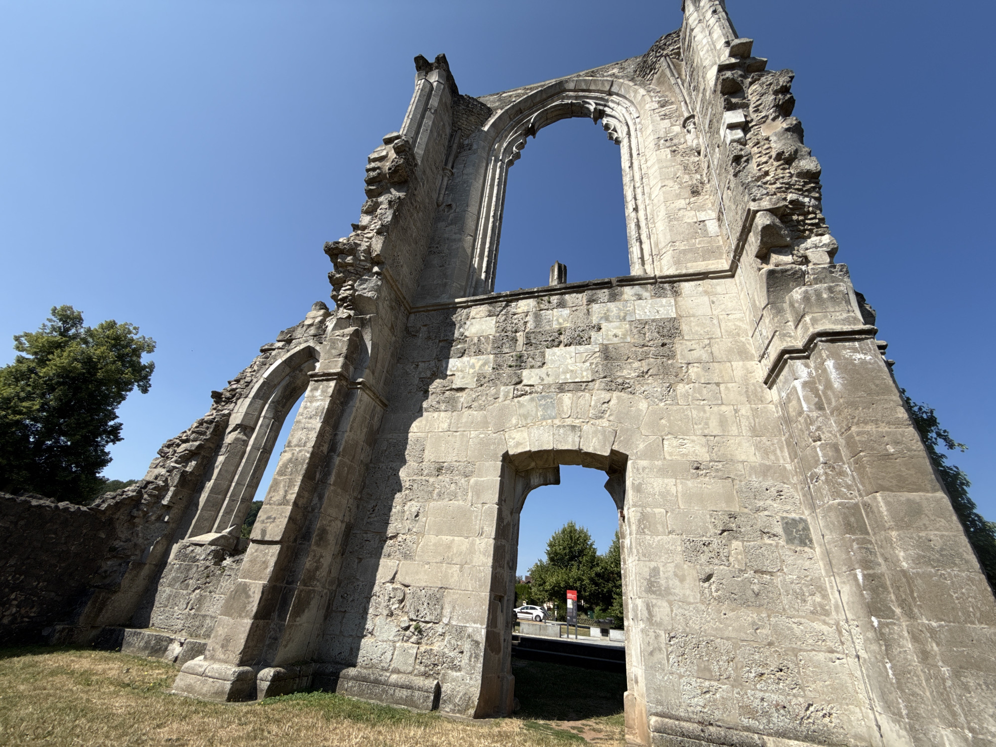 This is a low-angle, wide shot of the partially ruined facade of a Gothic-style stone structure, likely an abbey or church, under a clear blue sky. The stonework is aged and weathered, with some areas of collapse and missing sections. The main subject is the large, arched opening in the center of the facade, which offers a view of trees and a road with a white car. To the left, another, smaller arched window is visible, also framing a view of the exterior. The ground in the foreground is covered with dry grass. The structure is identified as the ruins of Walkenried Abbey, located in Walkenried, Germany.