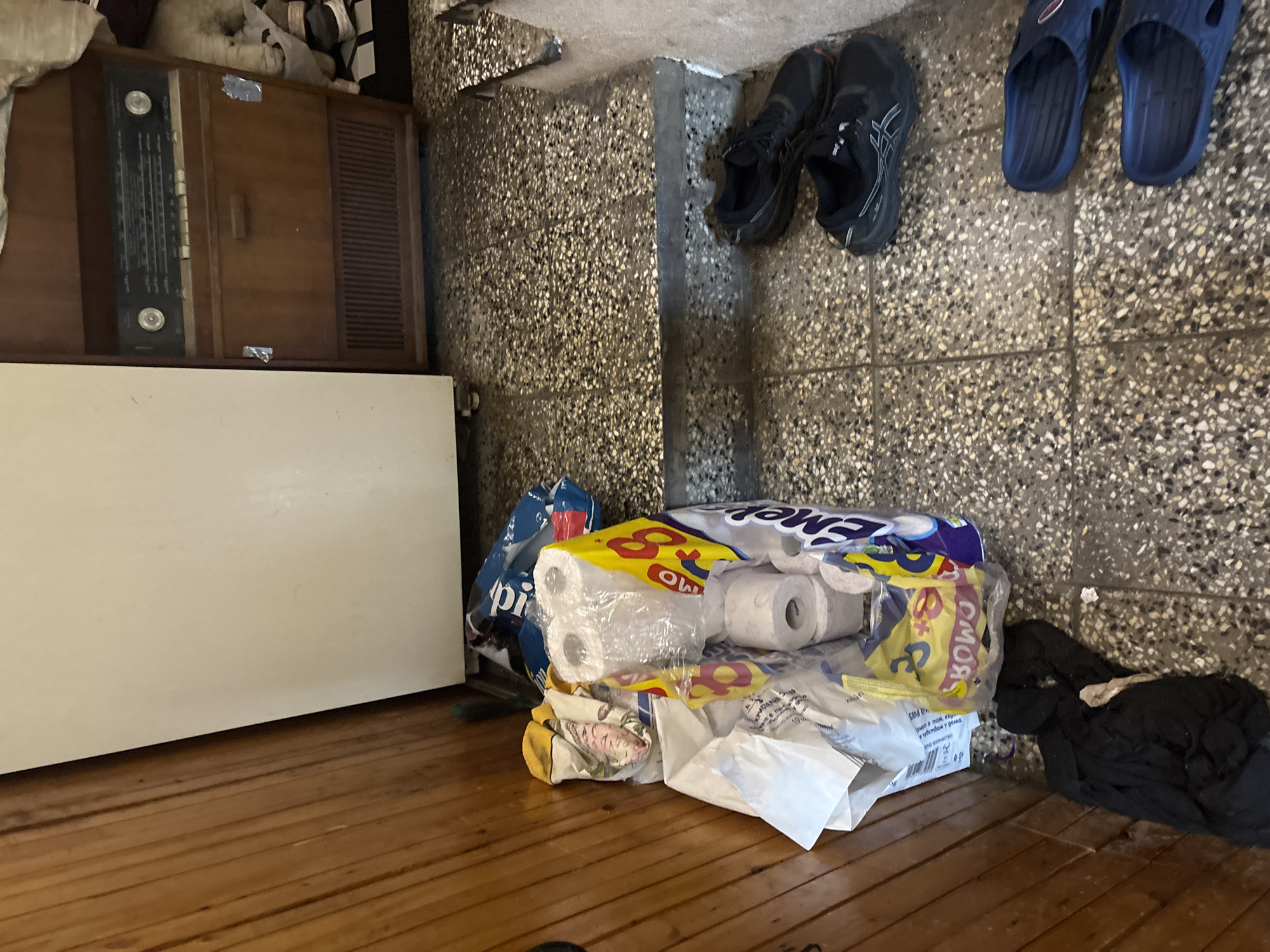 An indoor scene is depicted, focusing on a cluttered corner of a room. The floor is made of polished wooden planks, and the lower portion of the wall consists of speckled, light grey and dark grey tiles, typical of terrazzo.
On the left, a vintage-style wooden console unit with an integrated radio tuner and speaker grille is visible. The radio tuner panel is black with silver knobs and various frequency markings. Above and behind this unit, some folded fabrics or blankets are stacked. Next to the radio unit, a large, plain white rectangular object stands, possibly a refrigerator or a tall cabinet.
In the center and right foreground, numerous household items are piled on the wooden floor and against the tiled wall. These include several large plastic packages of toilet paper, one of which clearly shows the brand "OMO" and "Emer" (or part of a brand name) with some Cyrillic text including "чувствам у дома" (feeling at home) and "уте колчество" (comfort). Other plastic bags and possibly detergent packaging are also present.
A pair of black sneakers with white soles and subtle branding is resting near the tiled wall. Further right, a pair of blue plastic sandals or slides is placed on the tiled floor. Next to the piled packages, a heap of dark-colored clothing or fabric lies on the floor.
The lighting appears natural, suggesting daytime. The overall impression is that of a utility space or a storage area within a residence. No people are visible in the image.