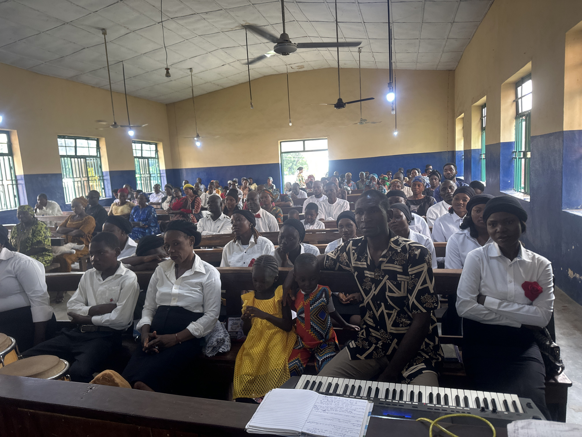 The image shows a congregation seated in a room with yellow walls and a white, tiled ceiling. Ceiling fans are visible, as are light bulbs hanging from wires. Windows with green bars are set high on the walls. In the foreground, a keyboard is placed on a wooden surface. Several people are seated on wooden pews, many of them wearing white shirts. Two young children, a girl in a yellow dress and a boy in patterned clothing, are seated in the front row. A woman in a white shirt and black skirt is seated to the right of the children, with her hands clasped. A man in a patterned shirt is seated to the left of the children. The overall scene suggests a gathering in a place of worship or a community hall. The location is identified as the city of Akwanga, Nigeria.
