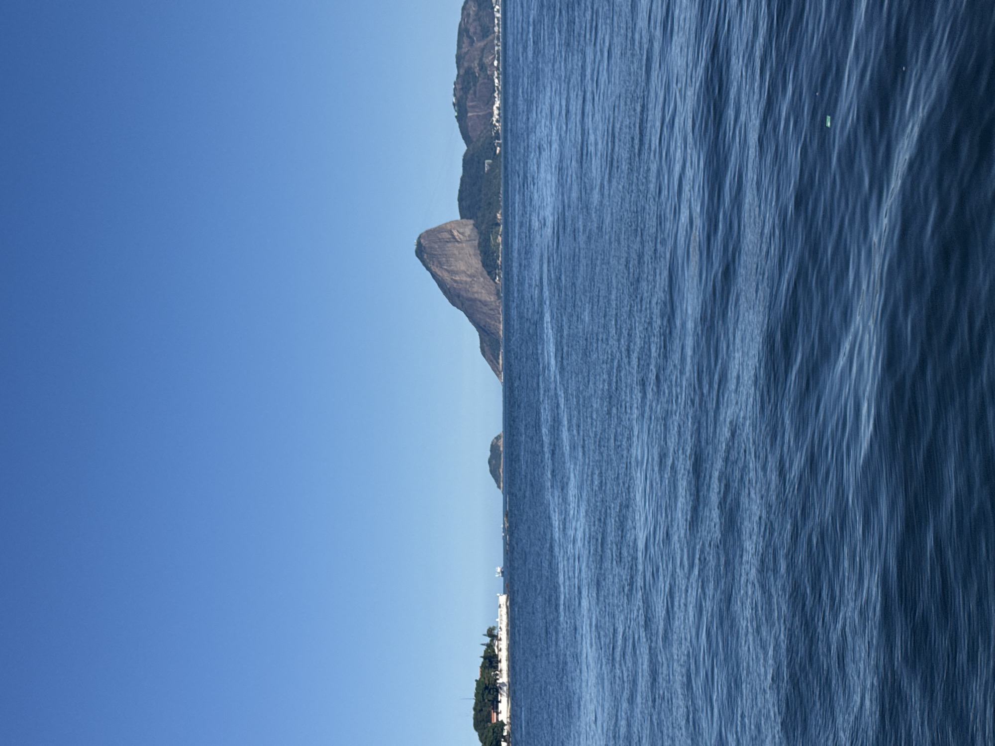 The image depicts a coastal scene with a clear blue sky on the left and the ocean on the right. The horizon line is not horizontal, suggesting the photo may have been taken from a boat or at an angle.
On the land side, the image shows a rugged, mountainous terrain covered in lush green vegetation. Several buildings, likely part of a town or city, are visible nestled on the slopes of the mountains. The architecture suggests a Mediterranean or tropical influence. A prominent rock formation juts out from the mountainside, adding to the dramatic landscape. In the foreground on the land side, there are trees and some white structures, possibly buildings or recreational facilities.
The ocean occupies the right side of the image and appears to be calm with gentle ripples. The water is a deep blue color, reflecting the clear sky. There are a few small, unidentifiable objects floating on the water's surface, appearing as small bright specks.
The overall impression is a scenic view of a coastal city or town bordering a natural mountainous landscape and the sea. The bright sky and calm water suggest it is daytime with fair weather. The location context of Niterói, Brazil, aligns with the visual elements of mountainous coastlines and proximity to the ocean. No people are discernible in the image. There is no visible text.