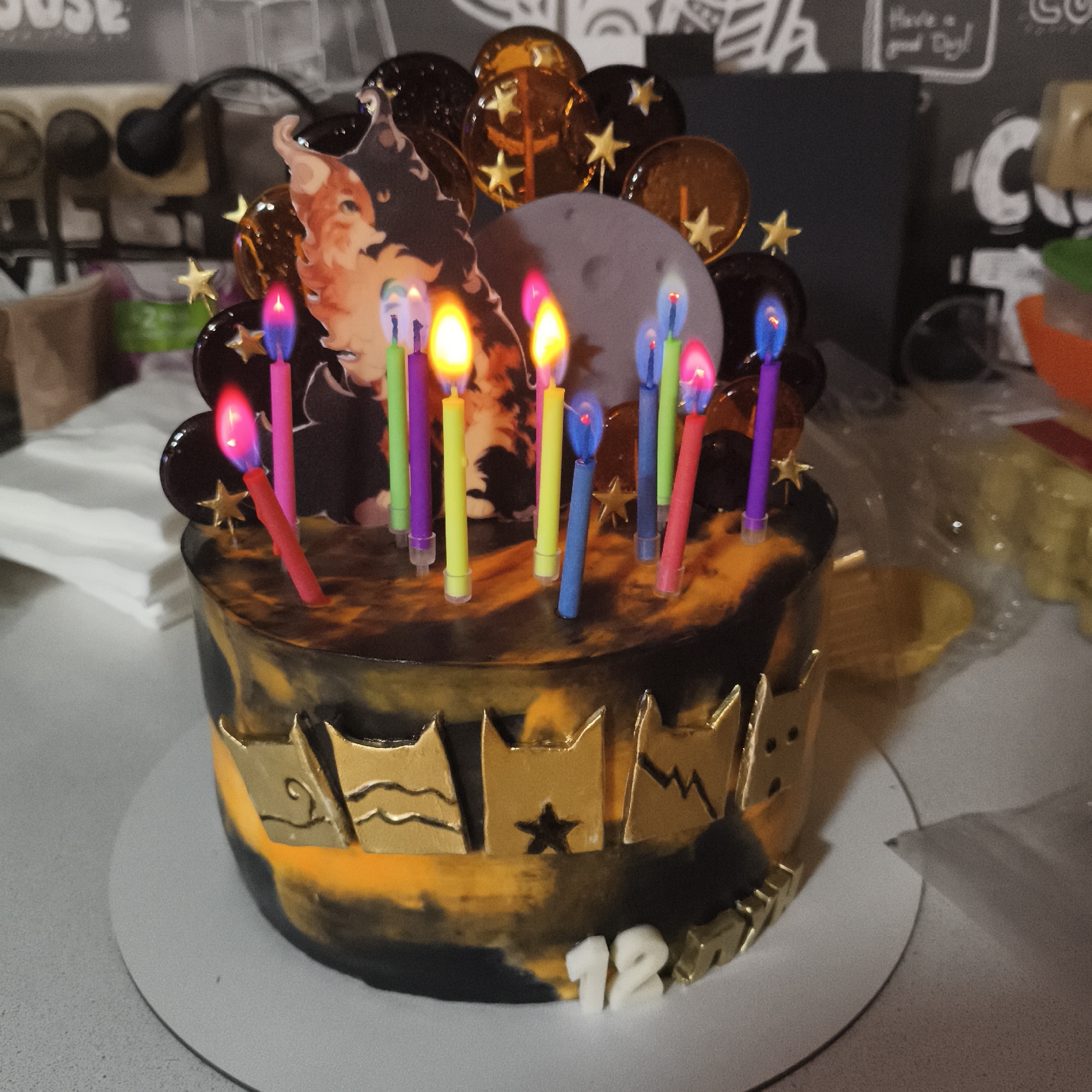 The image shows a decorated birthday cake with 12 lit candles on a white round base. The cake has a black and orange marbled frosting. Gold decorations adorn the cake including cat crown images and the number "12" at the base. Behind the candles, there is a depiction of an anime character, lollipops with stars, and a gray moon-shaped decoration. The cake is on a white surface in what appears to be a domestic kitchen setting, with a kitchen counter in the background with various kitchen utensils. I am unable to confirm the specific location in Chaikovsky, Russia, because I lack the ability to ascertain a physical location based on the image.