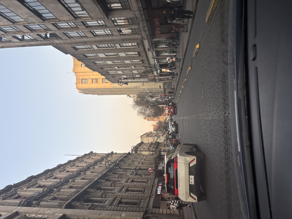 A street scene with tall buildings lining both sides. The buildings are predominantly gray with many windows. A street paved with bricks stretches between them, filled with cars and pedestrians. A white SUV with a visible license plate is in the foreground. The sky is visible between the buildings, transitioning from light blue to a faint orange near the horizon. The photo is taken from inside a vehicle.