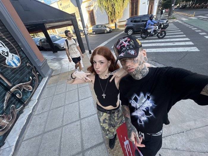 The image captures a wide-angle, slightly low-angle selfie or group shot featuring two individuals with distinctive styles, standing on a sidewalk in an urban setting. The time of day appears to be late afternoon or early evening, indicated by the soft, diffused sunlight.
In the foreground, on the right, is a man with extensive tattoos covering his face, neck, arms, and hands. He has large ear gauges and is wearing a black baseball cap with colorful patches and designs. He is dressed in a black oversized t-shirt featuring a prominent white and blue graphic design on the front. He holds a red rectangular object, possibly a sign or a board, with a large white letter "E" visible on it. His expression is neutral or serious as he looks directly at the camera.
Standing to his left, slightly behind him, is a young woman with reddish-brown hair, a light complexion, and freckles. She has a small nose ring and a tattoo on her left forearm. She is wearing a black sleeveless crop top, a silver cross necklace, and camouflage-patterned cargo pants with a dark belt. Her left hand is raised, forming a 'peace' or 'rock-on' gesture with her fingers, while her right hand rests lightly on the man's shoulder. She is also looking towards the camera with a slightly pouting expression.
The subjects are standing on a paved sidewalk composed of square tiles. To the immediate left of the woman, a building facade with an intricate dark-colored metal grate or decorative panel is visible. A blue and white striped oval shape, possibly part of a logo or artwork, is painted on this panel. Above this, a dark awning extends over what appears to be a storefront.
In the mid-ground, further down the sidewalk, another young man is walking away from the camera. He has dark hair, is wearing a light-colored t-shirt with the word "GENUINE" printed on it, black shorts, and headphones around his neck. Behind him, several cars are parked along the street.
In the background, on the street itself, a person on a motorcycle, wearing a blue jacket and a helmet, is crossing a white-striped pedestrian crosswalk. To the left of the motorcycle, a dark grey SUV (resembling a Jeep Renegade) is parked. Buildings with light-colored facades and various architectural details line the street in the distance. The name "ZHOUE" is visible on a sign on one of the buildings in the upper left background. Trees are scattered along the street, and the sky is bright and clear.