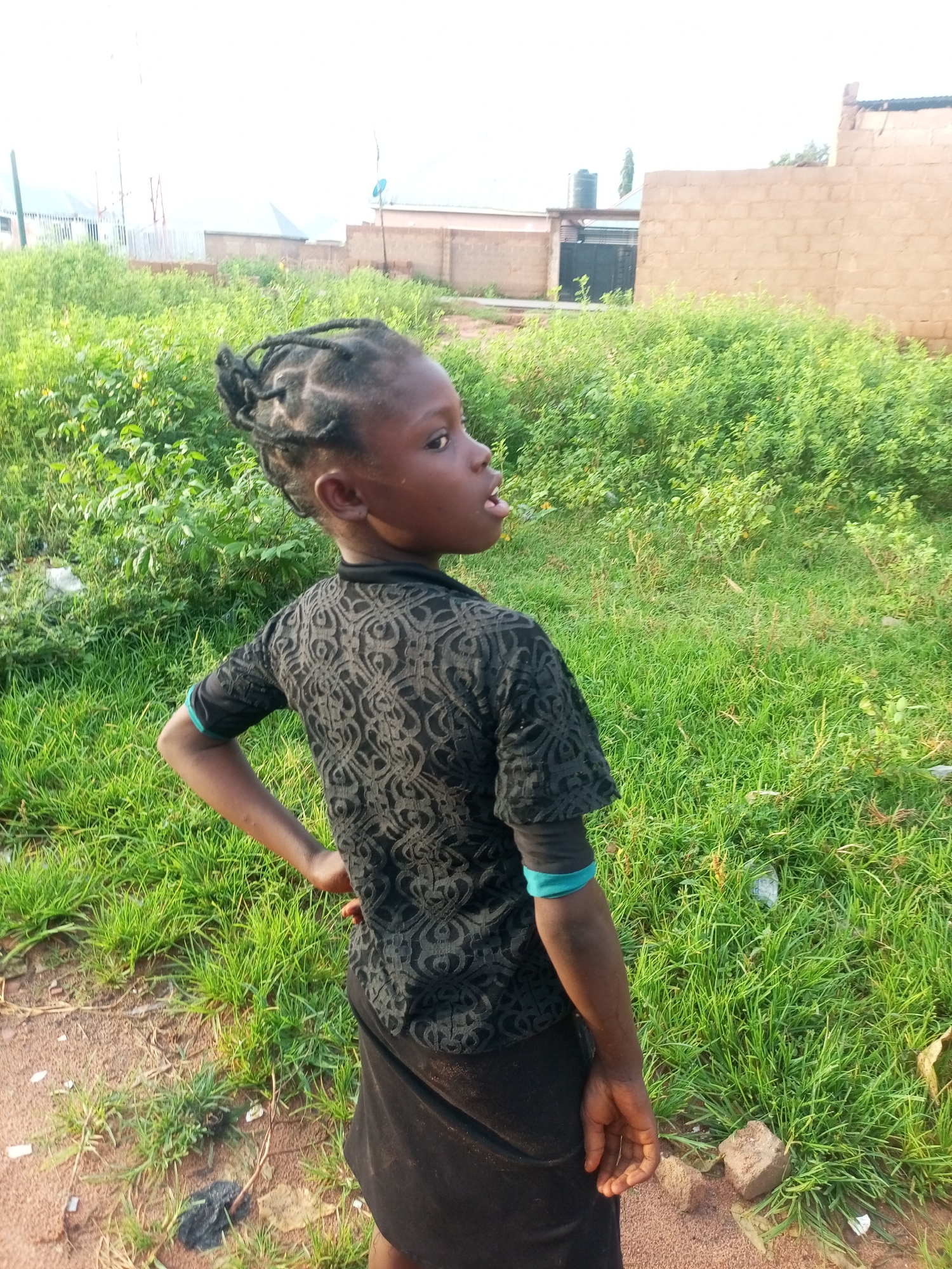 The image depicts a young Black girl with braided hair, wearing a black shirt and black skirt, standing outdoors in a grassy area. She is looking to her right with her tongue sticking out. In the background, there are buildings constructed from brown bricks and a large green water tank. The ground is a mixture of dirt and grass, with some scattered debris. The scene appears to be in a residential area, possibly in Tempure, Nigeria, given the architectural style and vegetation. The lighting suggests it is daytime.