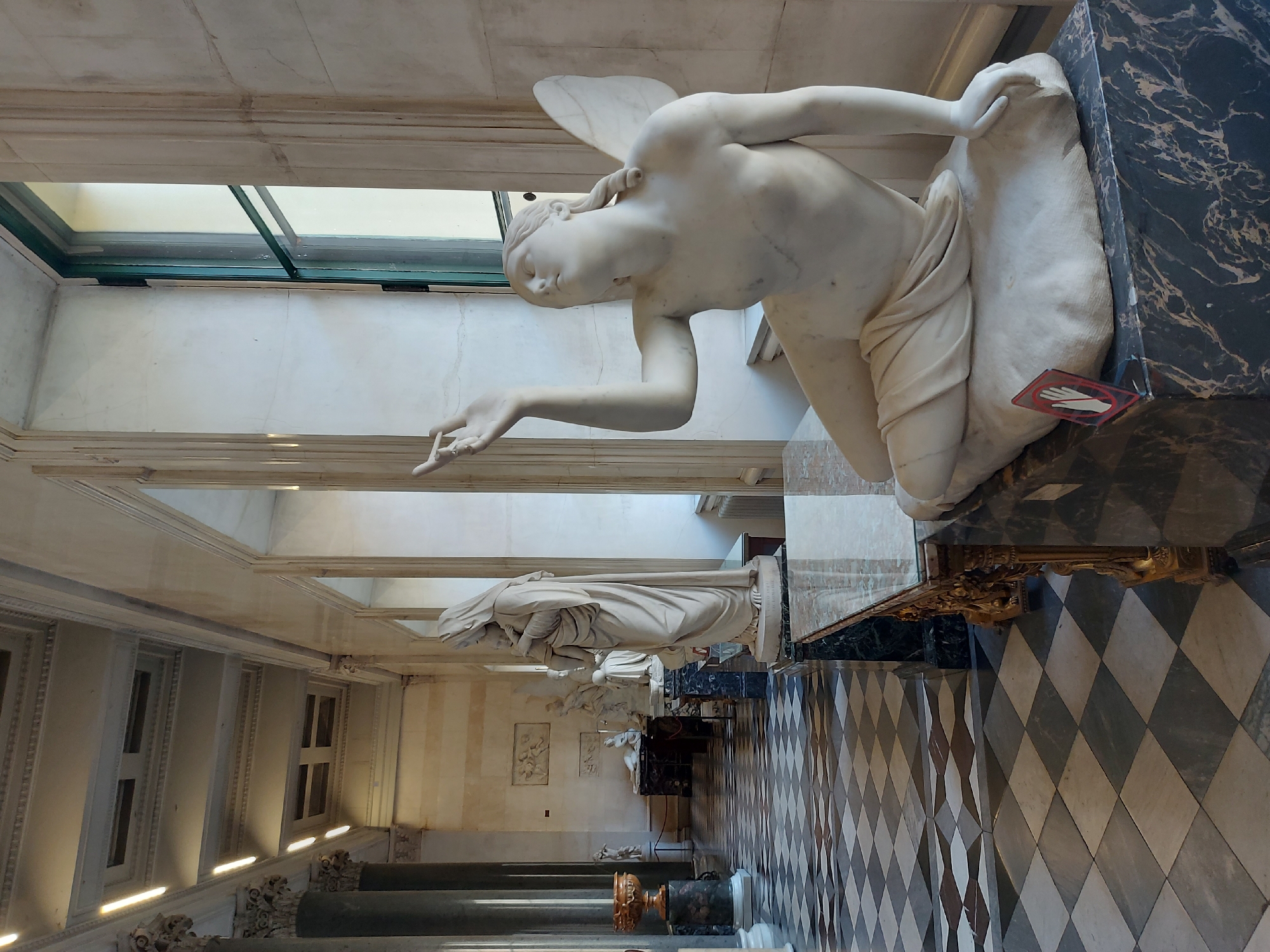 The image shows a gallery of sculptures in Saint Petersburg, Russia. The main subject is a white marble statue of a kneeling winged figure, likely an angel, positioned on a dark, veined marble base. Further down the gallery, additional white marble sculptures are visible. The floor is tiled in a black-and-white diamond pattern. The walls are white with windows on the left side providing natural light. Ornate columns and architectural details are present along the walls. A small, red sign with a white hand indicates "do not touch."