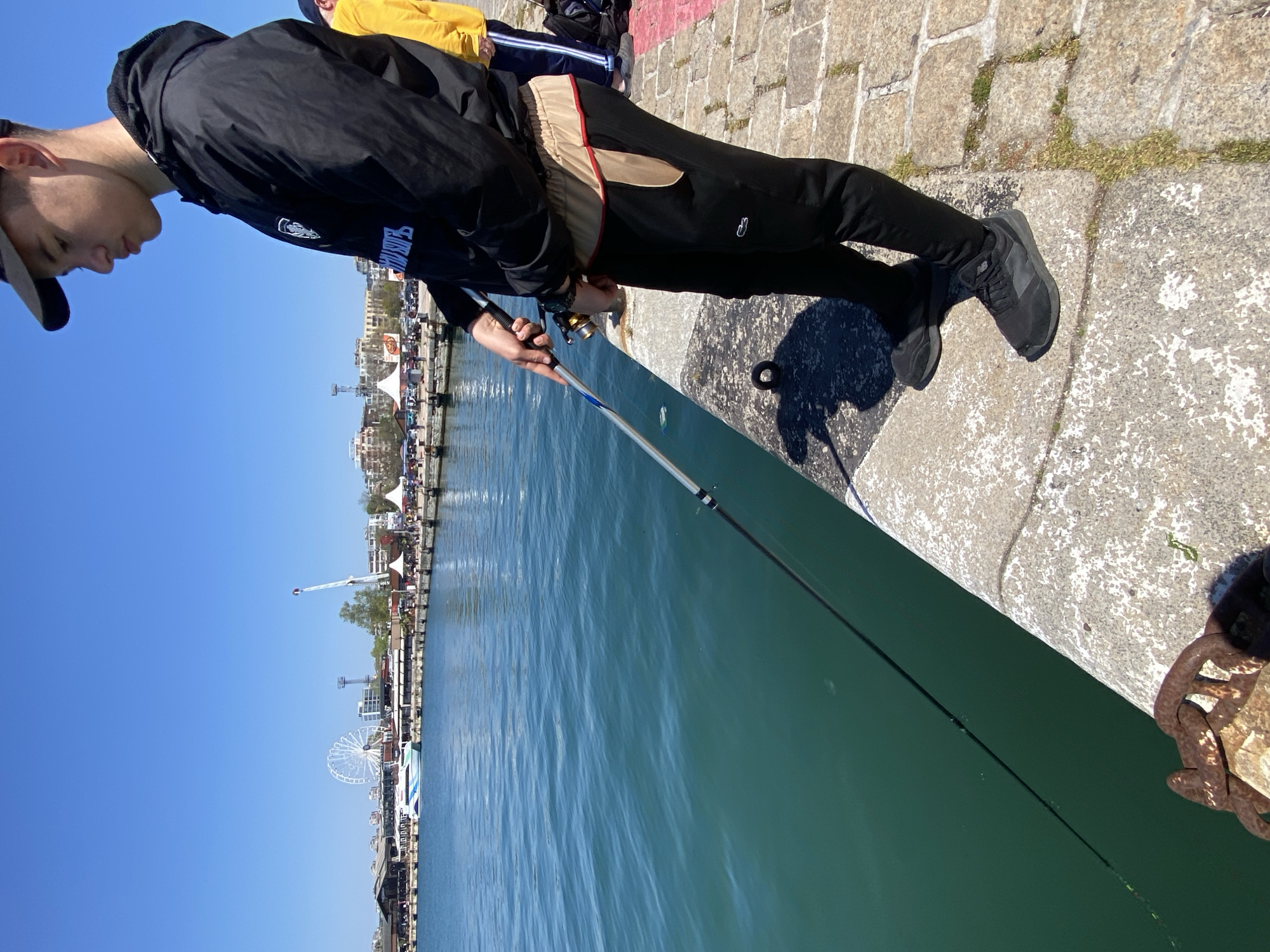 A young man is fishing from a concrete pier or quay. He is wearing a black jacket, black pants with tan and red trim, and black shoes. He holds a fishing rod over the edge, with the line extending into the water. Behind him, other people stand along the pier. In the background, there is a cityscape with a Ferris wheel and other structures, suggesting a location near a harbor or waterfront area. Based on the visual elements, this is likely the city of Varna, Bulgaria, specifically near the port or along the coast.