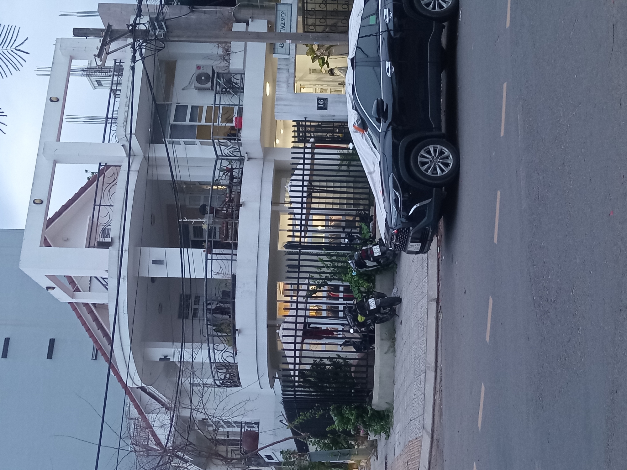 This media file captures a street scene in Đà Nẵng, Vietnam, likely during daytime given the natural light. The image is oriented vertically, showing a multi-story building with white balconies and a dark-colored car parked alongside the road. The building appears to be a residential or commercial space, with several windows and what looks like an air conditioning unit visible. A black iron fence runs along the sidewalk in front of the building, with some greenery and several parked motorcycles behind it.
The car, a dark SUV, is partially covered with a white sheet. Its wheels and part of its body are visible. A number plate is discernible on the car. The street itself is asphalt with yellow lane markings. To the left of the car, a sidewalk made of paving stones is visible, bordered by a small strip of grass. Further left, another building facade is visible, painted in a light blue hue. Overhead, electrical wires and poles are prominent, suggesting urban infrastructure.
A sign with the number "16" is attached to the building, and another sign with faded text that appears to read "STAGE SHOP" is also visible. The overall impression is that of a typical street in a Vietnamese city, with a mix of residential and commercial elements, and everyday vehicles. There are no clearly discernible people in the image, but the presence of parked motorcycles suggests recent activity. The weather appears to be overcast or hazy, as there are no strong shadows.