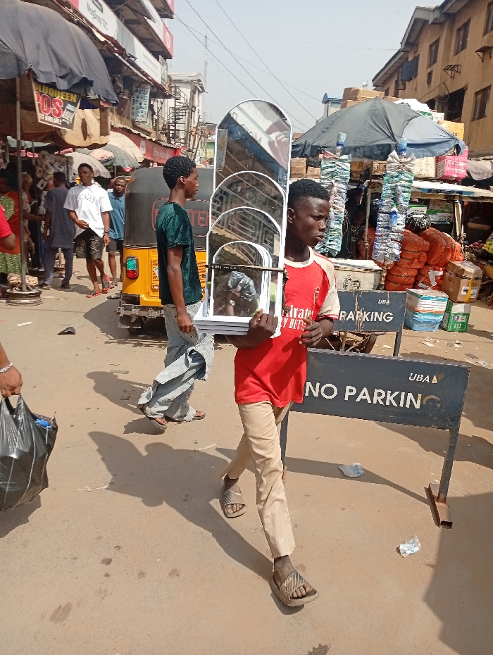 Two young men walk down a busy street. One wears a green shirt and the other a red shirt and tan pants. The red-shirted youth carries four arched mirrors, one in front of the other. Behind them is a yellow auto rickshaw. A "NO PARKING" sign is visible in the foreground. The street is crowded with pedestrians and vendors under umbrellas, creating a bustling marketplace atmosphere. Buildings and power lines are visible in the background under a hazy sky.