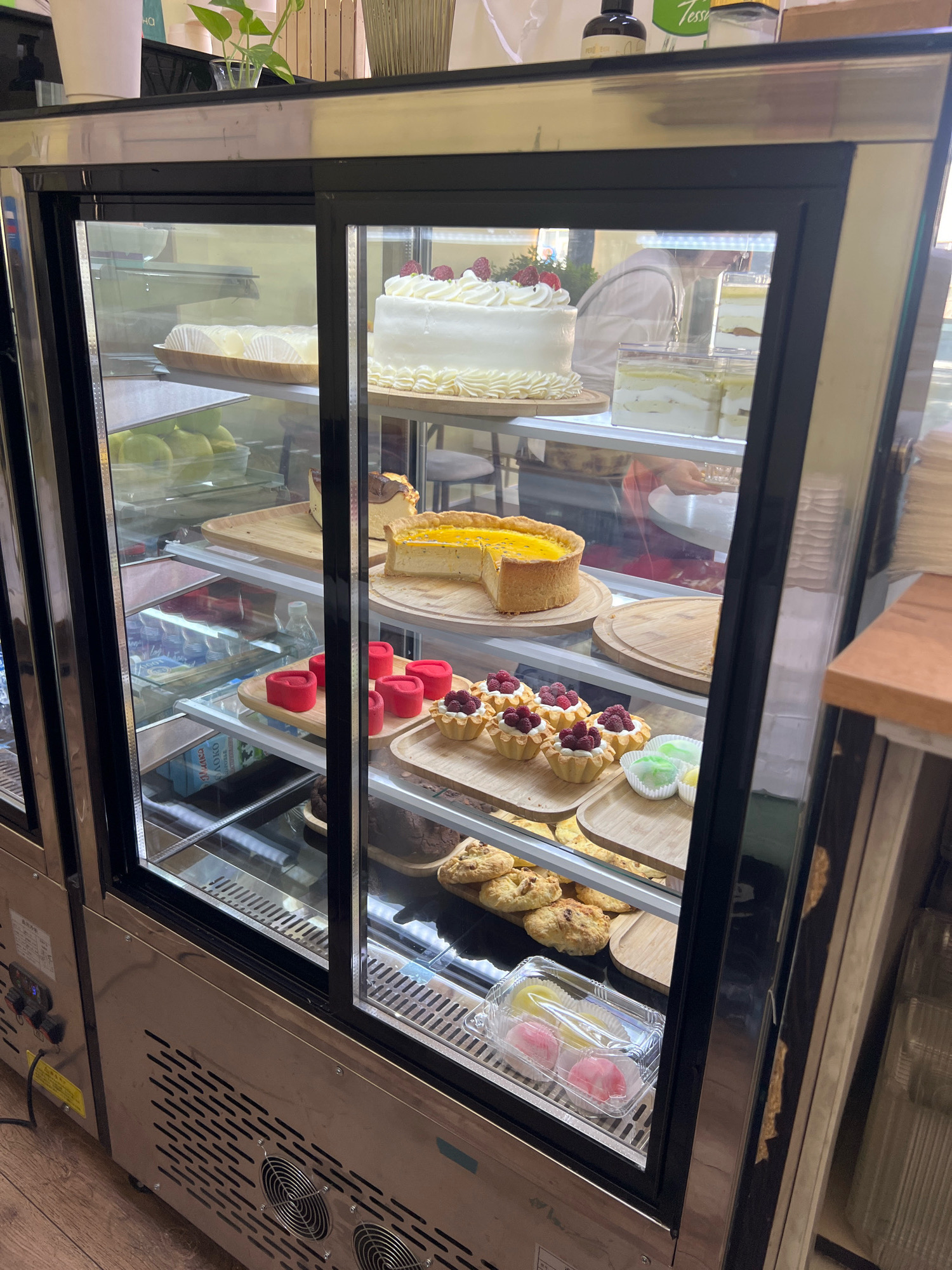 A commercial refrigerated display case filled with various desserts is shown in a store. The display case has glass doors and shelves made of wood and glass. Among the desserts are a white cake with raspberries, fruit tarts, a lemon tart, and mochi. On the shelves below, there are also some green fruits and pastries. In the background, a person wearing a light-colored shirt is visible. The exterior of the display case is stainless steel with vents and control panels. The floor appears to be made of wood. The location is identified as Osh City, Kyrgyzstan.