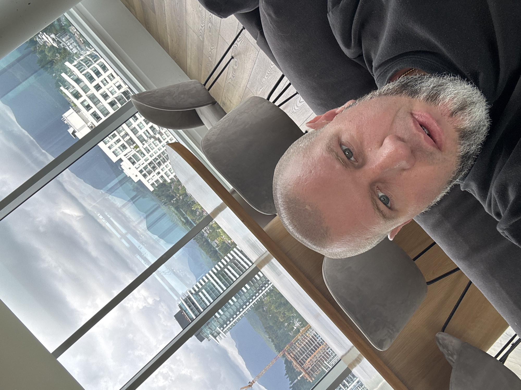A close-up selfie taken from a high vantage point in a modern apartment or office, overlooking a cityscape with mountains in the background. A bald, middle-aged Caucasian male with a grey beard is the primary subject, looking directly at the camera with a neutral expression. He is wearing a black crewneck sweatshirt. The background reveals a large window with multiple panes, offering a panoramic view of Coquitlam, Canada. Visible in the distance are several tall, modern white buildings, some appearing to be residential high-rises, and a construction site with a crane. The natural landscape features lush green trees and imposing mountains under a cloudy sky. Inside, modern grey upholstered chairs and a wooden table are partially visible, suggesting a contemporary interior design. The floor is made of polished wood planks. The lighting is natural, coming from the large windows.