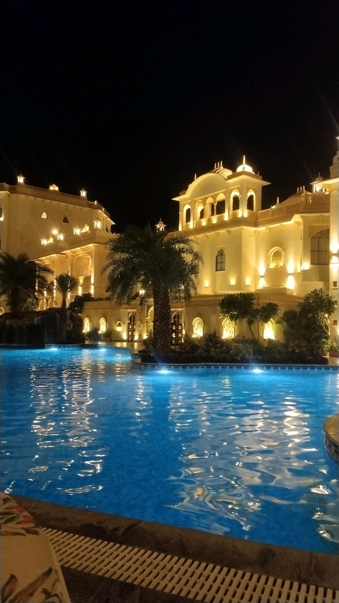 A photograph taken at night shows a swimming pool in front of a large, brightly lit building with ornate architecture, possibly a hotel or palace. The building is illuminated with warm yellow lights, highlighting its arched windows and decorative elements. Several palm trees are visible, one prominent in the foreground near the pool. The water in the pool reflects the lights of the building, creating a shimmering effect. The edge of the pool is visible in the foreground with a white grate. The sky is dark and clear. The image is likely taken in Munderi, India, given the architectural style and vegetation. No people are present in the image.