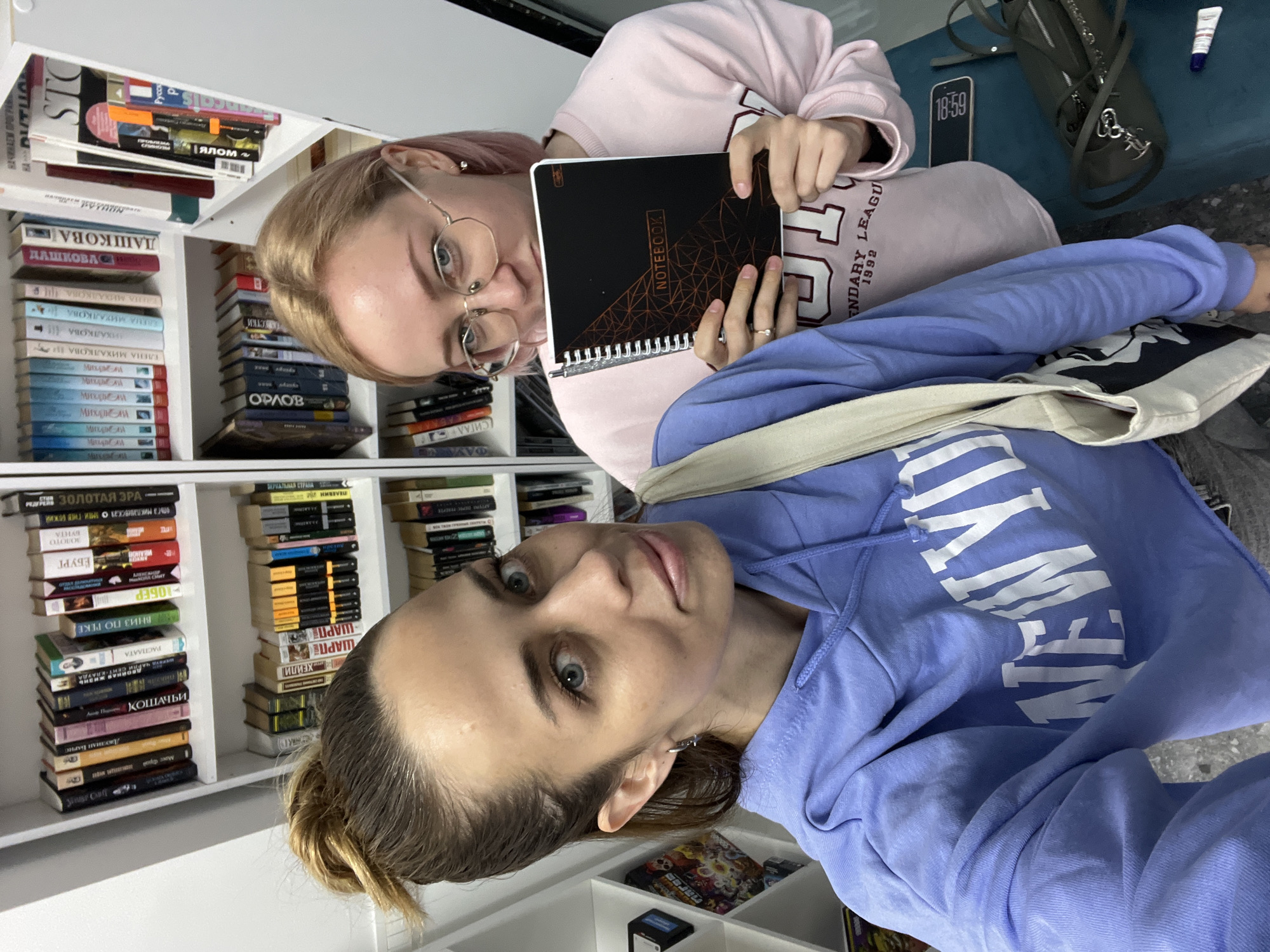 Two women are posing for a selfie in front of a bookshelf filled with various books, including many written in Russian. The woman in the foreground is wearing a light blue hoodie and has her hair in a bun. The woman in the background is wearing a pink sweatshirt and holding up a black notebook. A phone, handbag, and tube of cream are visible on a blue surface behind them. The environment appears to be indoors, potentially in a home or library. The specific location is not discernible.