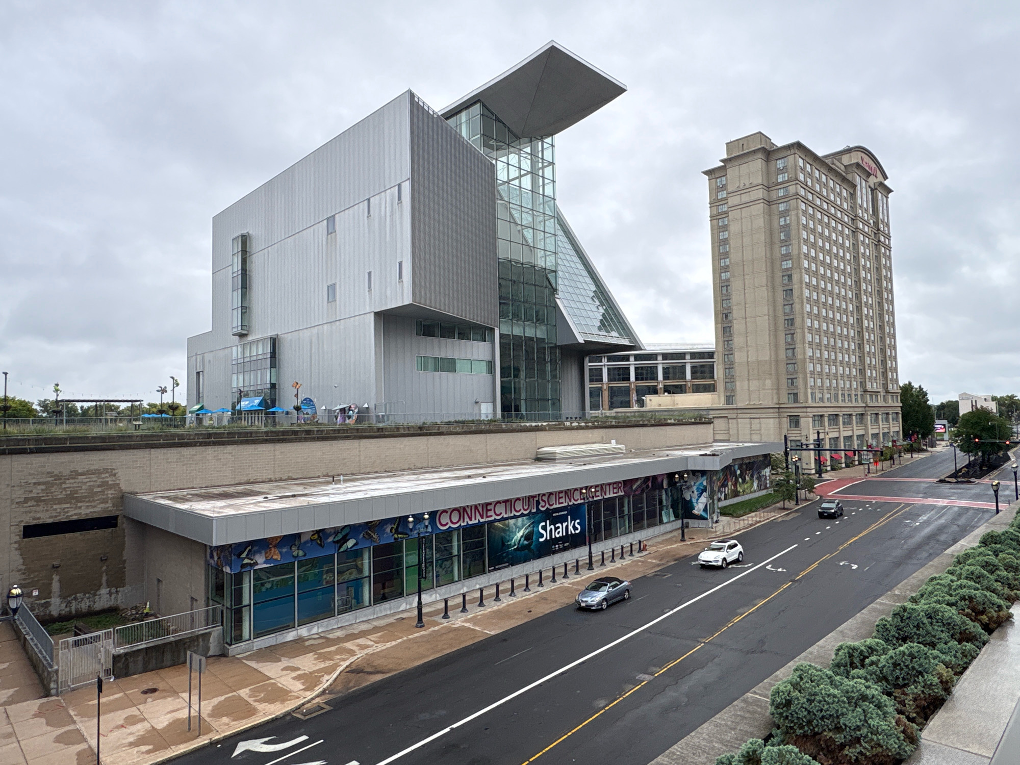 The image depicts the exterior of the Connecticut Science Center in Hartford, Connecticut, on a cloudy day. The science center is a modern, multi-story building with a distinctive angular design, featuring large glass sections and metallic siding. Adjacent to it stands a tall, beige high-rise building, possibly a hotel or office building, with numerous windows. A street runs in front of the science center, with a few cars visible. The street is wet, suggesting recent rain. On the right side of the image, a dense row of green bushes lines a raised area. The sky is overcast with gray clouds. The scene is captured from an elevated vantage point, looking down and across at the buildings and street.