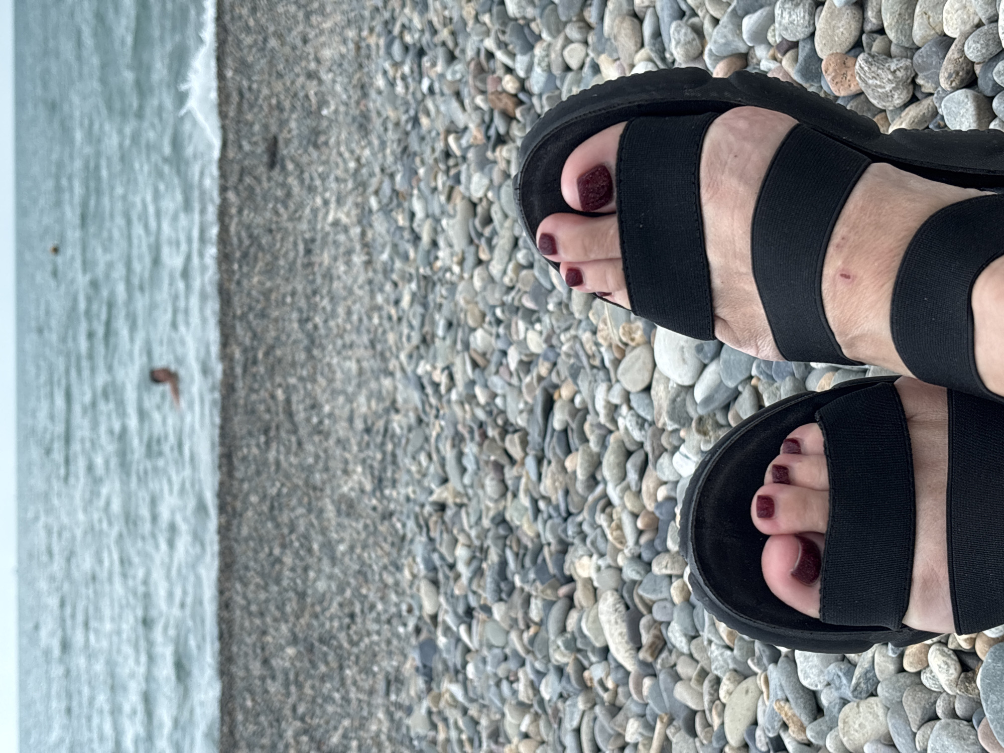 The media file depicts a close-up, low-angle shot of two feet wearing black elastic strap sandals, resting on a bed of grey and tan pebbles. The pebbles suggest a beach or coastal environment. To the left of the feet, a body of water is visible, appearing slightly choppy and a muted blue-grey color, indicative of a cool or overcast day. The water's edge is indistinct, blending with the pebble shore.
One foot shows toenails painted a deep maroon or burgundy color. A small, reddish abrasion is visible on the instep of the other foot, peeking out from the sandal straps. The lighting is soft and diffused, suggesting either an overcast sky or indirect sunlight, consistent with a cloudy day. There is no visible text within the image. The overall mood is calm and relaxed, capturing a moment of pause in a natural, seaside setting, possibly in Sochi, Russia, given the location context.