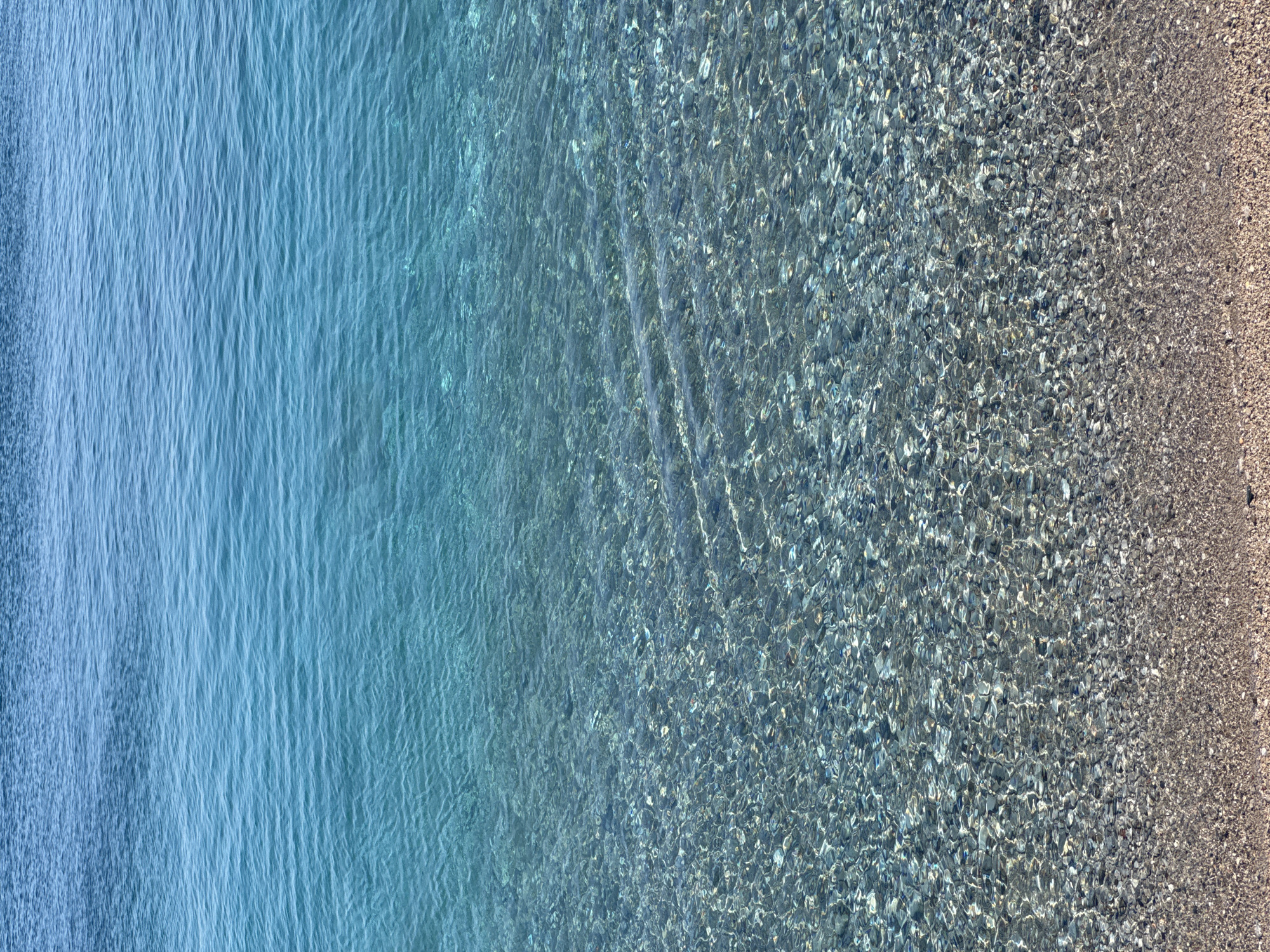 The image shows a top-down view of a shoreline. Clear blue water with subtle ripples transitions into a shallow area where the seabed is visible, revealing pebbles and stones. The right edge of the image shows a sandy beach. The location is identified as Prijevor, Montenegro, but no further information is present regarding the specific location. No people or actions are depicted.