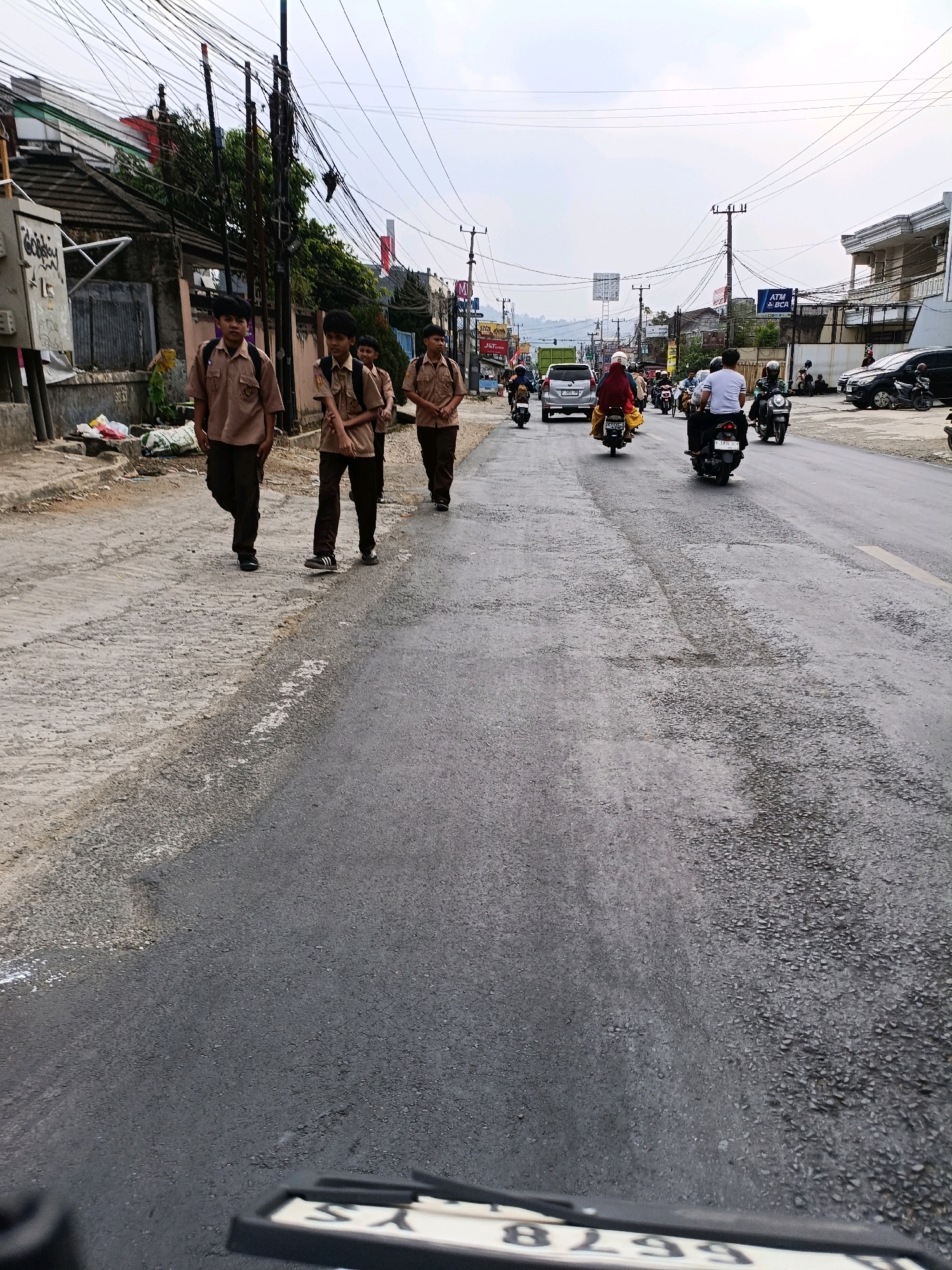 In Cibadak, Indonesia, three school-aged boys in identical brown uniforms and dark pants walk on the side of a paved road. They are carrying backpacks. The road is lined with various businesses and residential buildings. Motorcyclists and a car are visible on the road, indicating a moderately busy urban environment. Overhead, a complex network of electrical wires crisscrosses the sky. The road surface shows signs of wear and tear, with patches of asphalt missing. The weather appears overcast.