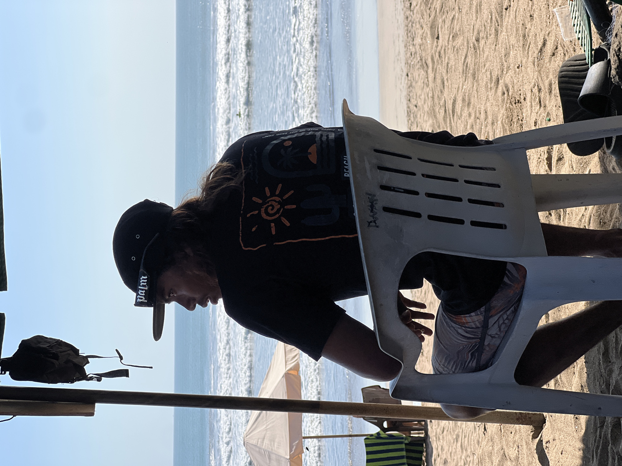 A medium shot captures a person sitting in a white plastic chair on a sandy beach in Kuta, Indonesia. The individual is viewed from behind and the side, wearing a black t-shirt with an orange graphic of a sun, cactus, and palm tree, and a black baseball cap with the word "palm" in white lettering. They are looking out towards the ocean, where waves are breaking. Behind the person, a beach umbrella and striped chairs are visible. The scene is illuminated by bright sunlight, casting shadows on the sand.