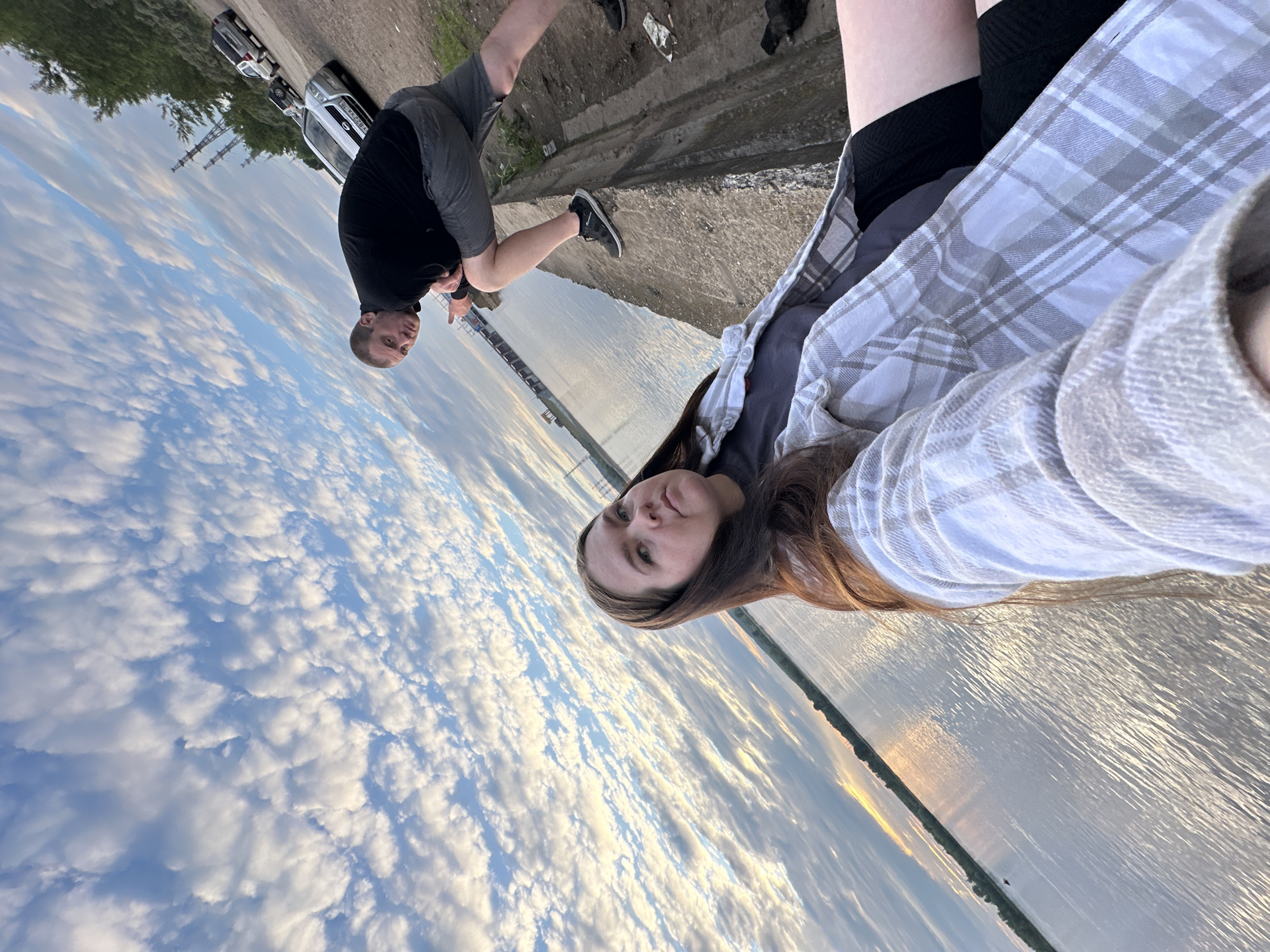 Two individuals, a woman in the foreground and a man in the background, are positioned on a concrete embankment overlooking a body of water. The woman is facing forward, her head turned towards the camera, and she is wearing a plaid shirt over a dark t-shirt and black shorts. The man is behind her, crouched down and looking towards the camera, wearing a black long-sleeved shirt and grey shorts. Several cars are parked on a dirt road behind the man, and industrial structures and trees are visible in the distance. The sky above is filled with scattered white clouds against a blue sky, with a hint of sunset light on the horizon. The water surface is rippled and reflects the light.
