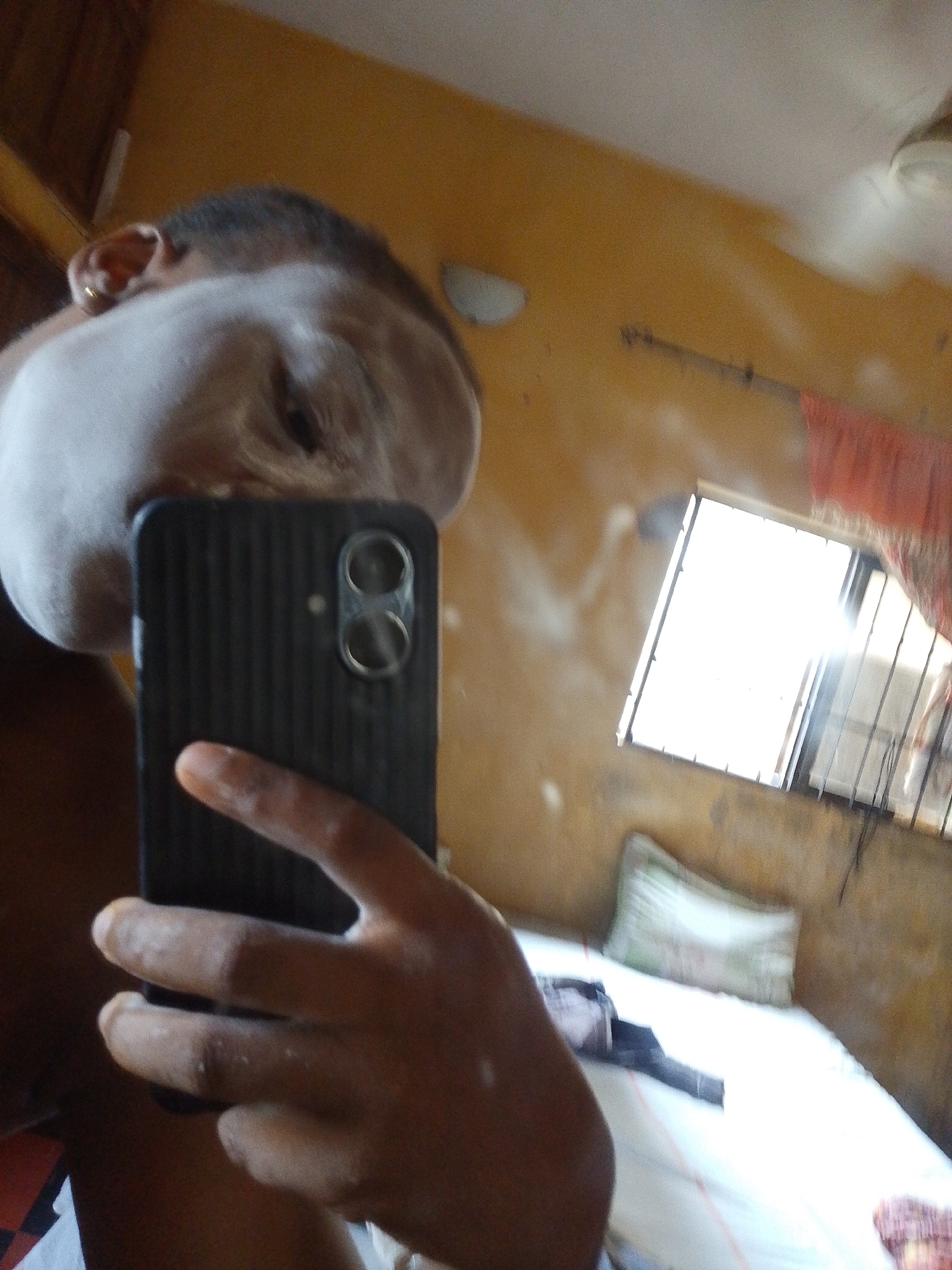 The image shows a person taking a selfie in a room. The person's face is partially obscured by the phone. Visible behind the person is a bed with white sheets, a pillow, and what appears to be clothing or a bag on it. A window with bars is in the background, letting in bright sunlight. The walls are painted yellow and appear to be somewhat worn. The image is taken in Umuahia, Nigeria.